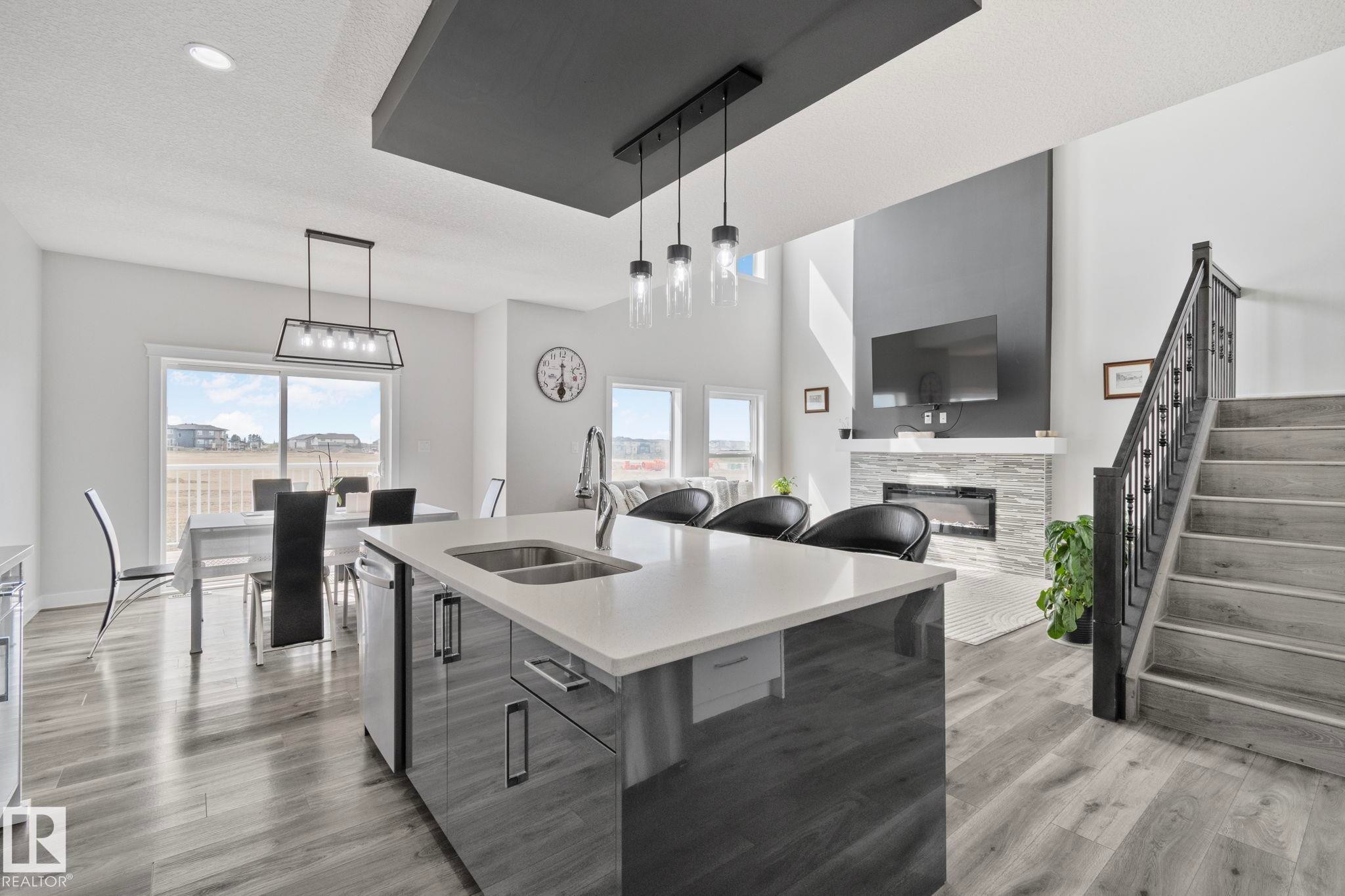 Open-concept interior featuring a kitchen island with a double sink and white countertop, wood-finish flooring, a contemporary fireplace with a stone surround, a prominent staircase with dark railings, and sliding glass doors leading to an exterior deck - 334 Aston Close, Leduc, AB - Indoor Photo Showing Kitchen With Double Sink With Upgraded Kitchen