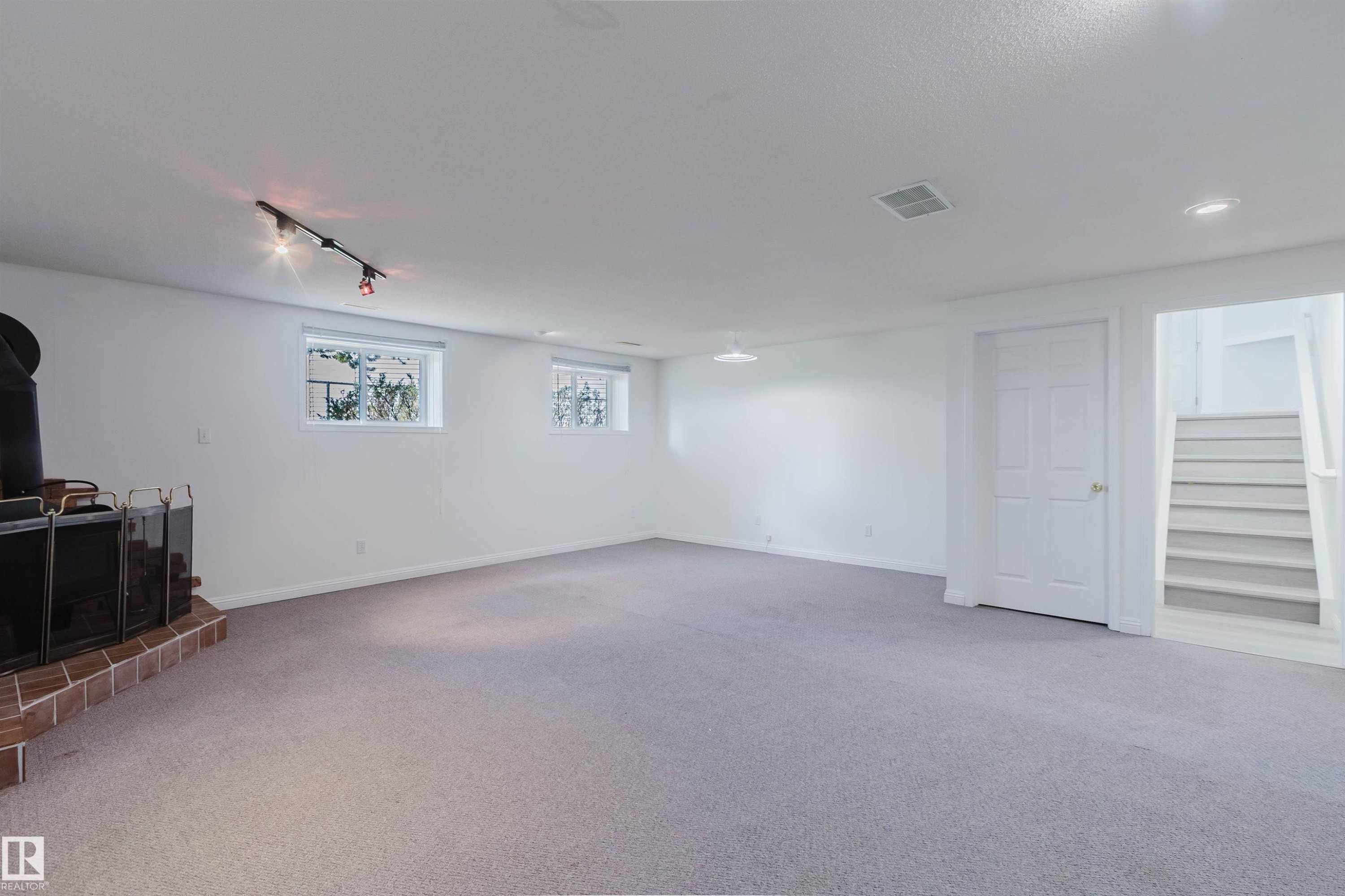 Spacious carpeted room featuring a freestanding stove with a brick hearth, multiple windows, a track lighting fixture, and a white interior door, with stairs visible beyond - 13755 131B Avenue, Edmonton, AB - Indoor Photo Showing Other Room