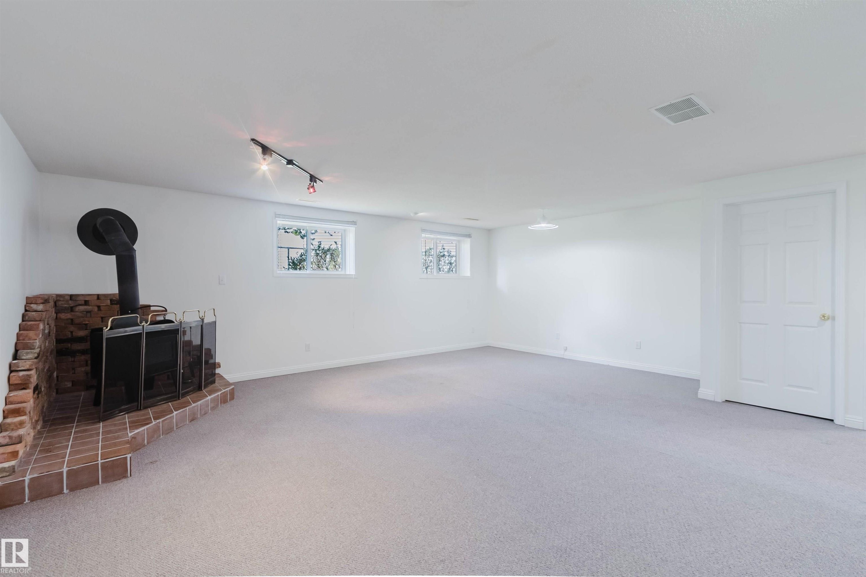Spacious room featuring a brick hearth and wood-burning stove, light-colored carpeting, white walls, two windows, and track lighting - 13755 131B Avenue, Edmonton, AB - Indoor Photo Showing Other Room With Fireplace