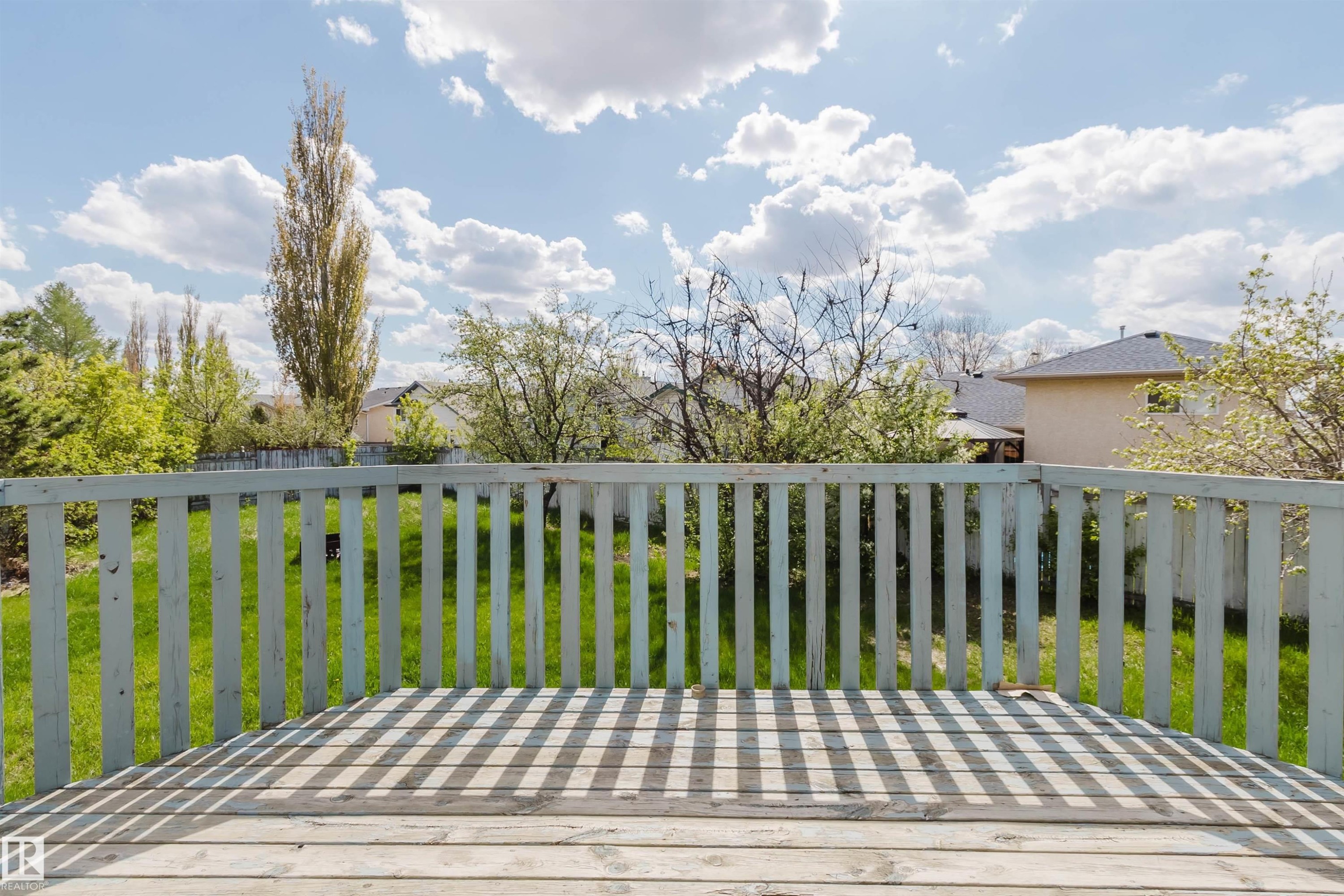 Spacious outdoor deck with a wooden railing and decking, overlooking a landscaped backyard with lush green grass and mature trees - 13755 131B Avenue, Edmonton, AB - Outdoor