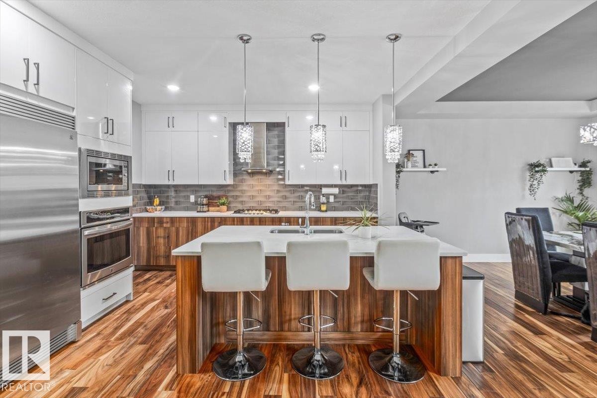 Modern kitchen featuring white cabinetry, stainless steel appliances, a center island with seating, a tile backsplash, and wood-finish flooring - 219 Cranberry Bend, Fort Saskatchewan, AB - Indoor Photo Showing Other Room