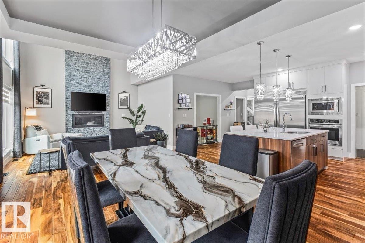 Open-concept living space featuring wood-finish flooring, a stone-accented fireplace, and a contemporary crystal chandelier - 219 Cranberry Bend, Fort Saskatchewan, AB - Indoor Photo Showing Dining Room