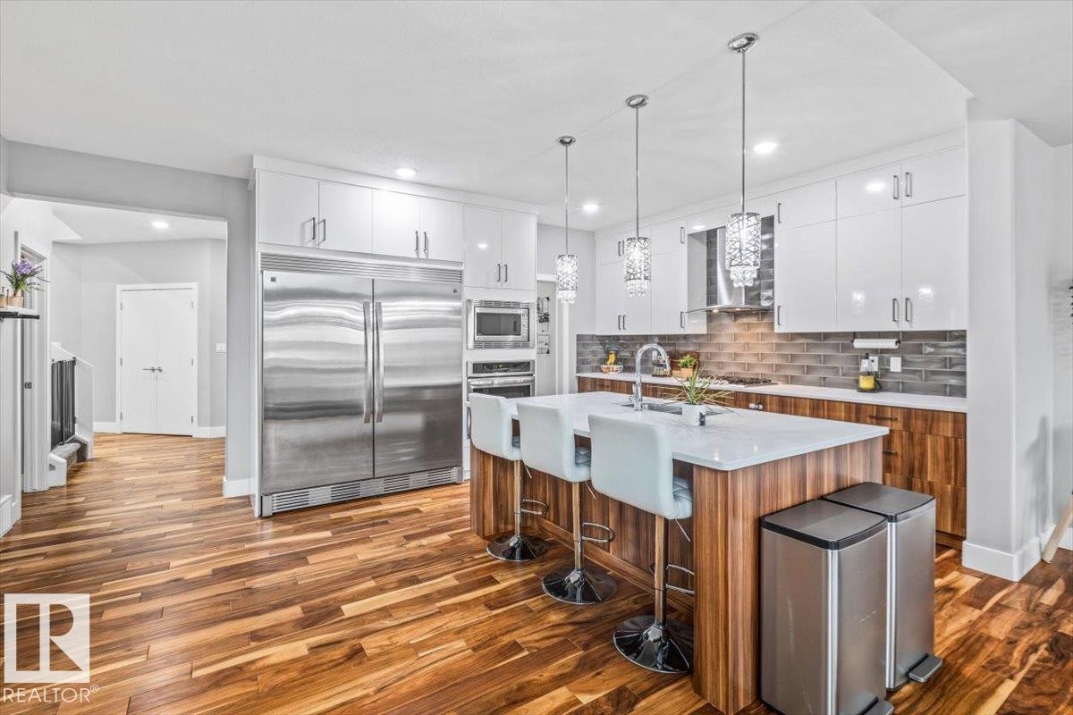 Modern kitchen featuring wood-finish flooring, white flat-panel cabinetry, stainless steel appliances, a center island with seating, and decorative pendant lighting - 219 Cranberry Bend, Fort Saskatchewan, AB - Indoor Photo Showing Kitchen With Stainless Steel Kitchen With Upgraded Kitchen