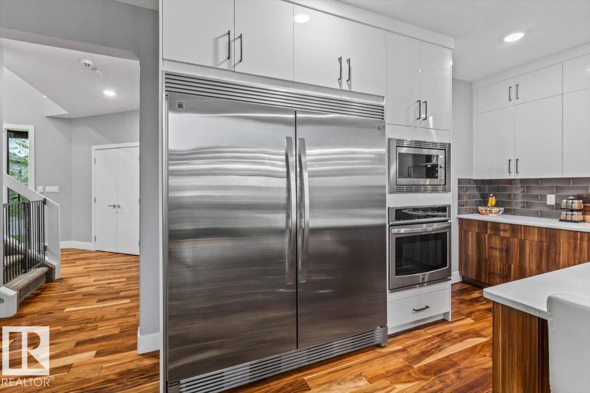 Gourmet kitchen featuring a large stainless steel refrigerator, built-in microwave, wall oven, glossy white upper cabinetry, and contrasting wood-finish lower cabinetry - 219 Cranberry Bend, Fort Saskatchewan, AB - Indoor Photo Showing Kitchen With Stainless Steel Kitchen With Upgraded Kitchen