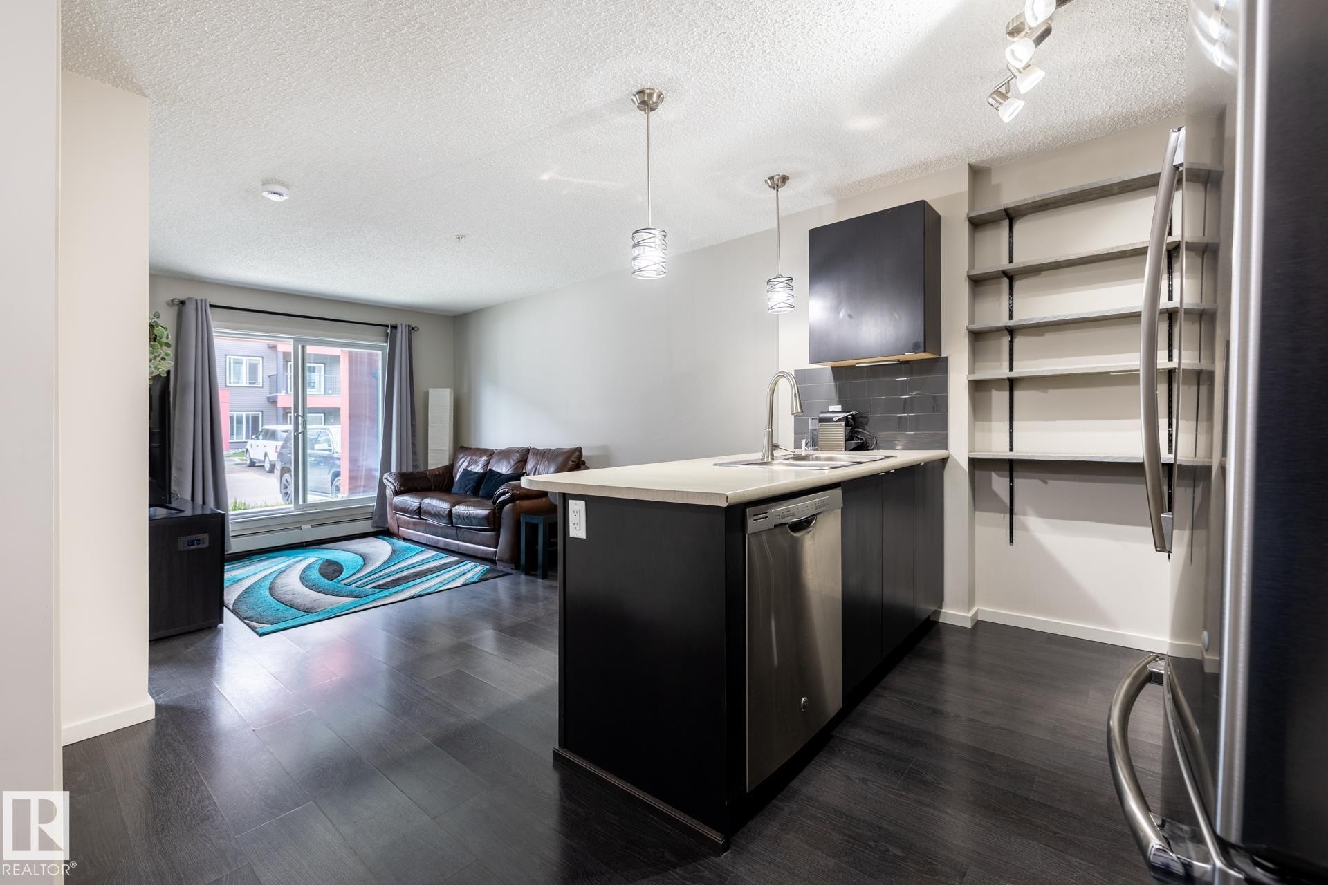 Open-concept living space featuring dark wood-finish flooring, a kitchen island with a light-toned countertop, stainless steel appliances, and built-in shelving - 131 344 Windermere Road, Edmonton, AB - Indoor Photo Showing Kitchen With Upgraded Kitchen