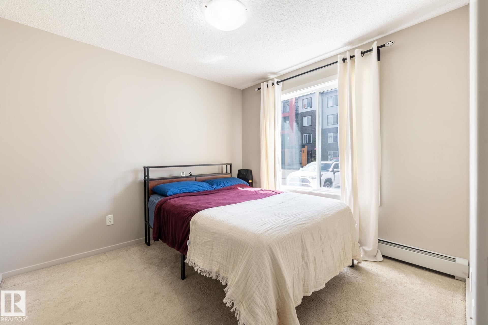 Bedroom featuring neutral wall paint, light-colored carpet, a large window with white curtains, and a ceiling-mounted light fixture - 131 344 Windermere Road, Edmonton, AB - Indoor Photo Showing Bedroom