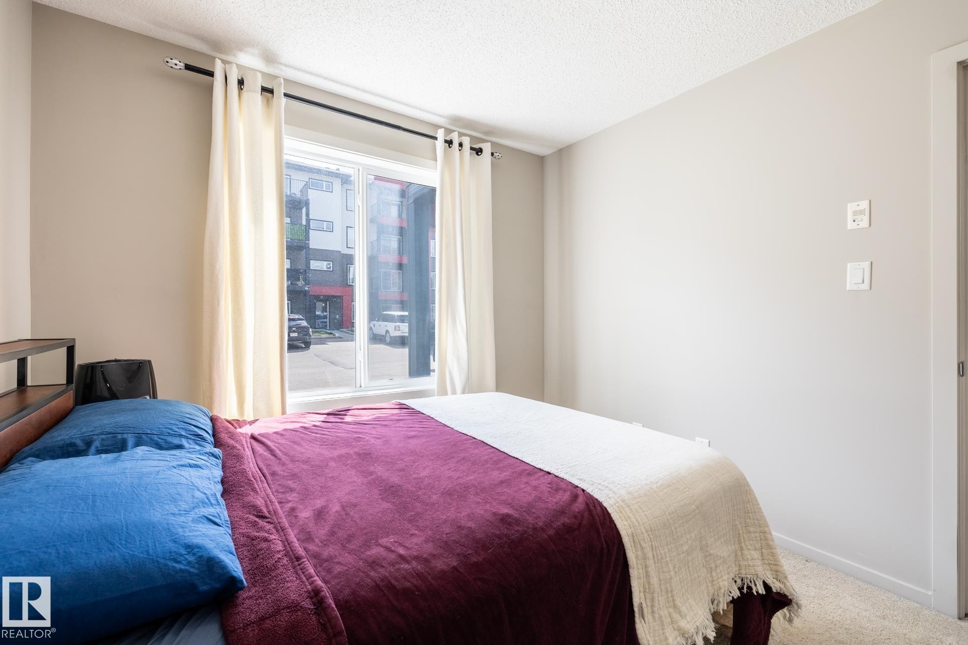 Bedroom featuring a large window, textured ceiling, light neutral wall paint, and soft carpet flooring - 131 344 Windermere Road, Edmonton, AB - Indoor Photo Showing Bedroom