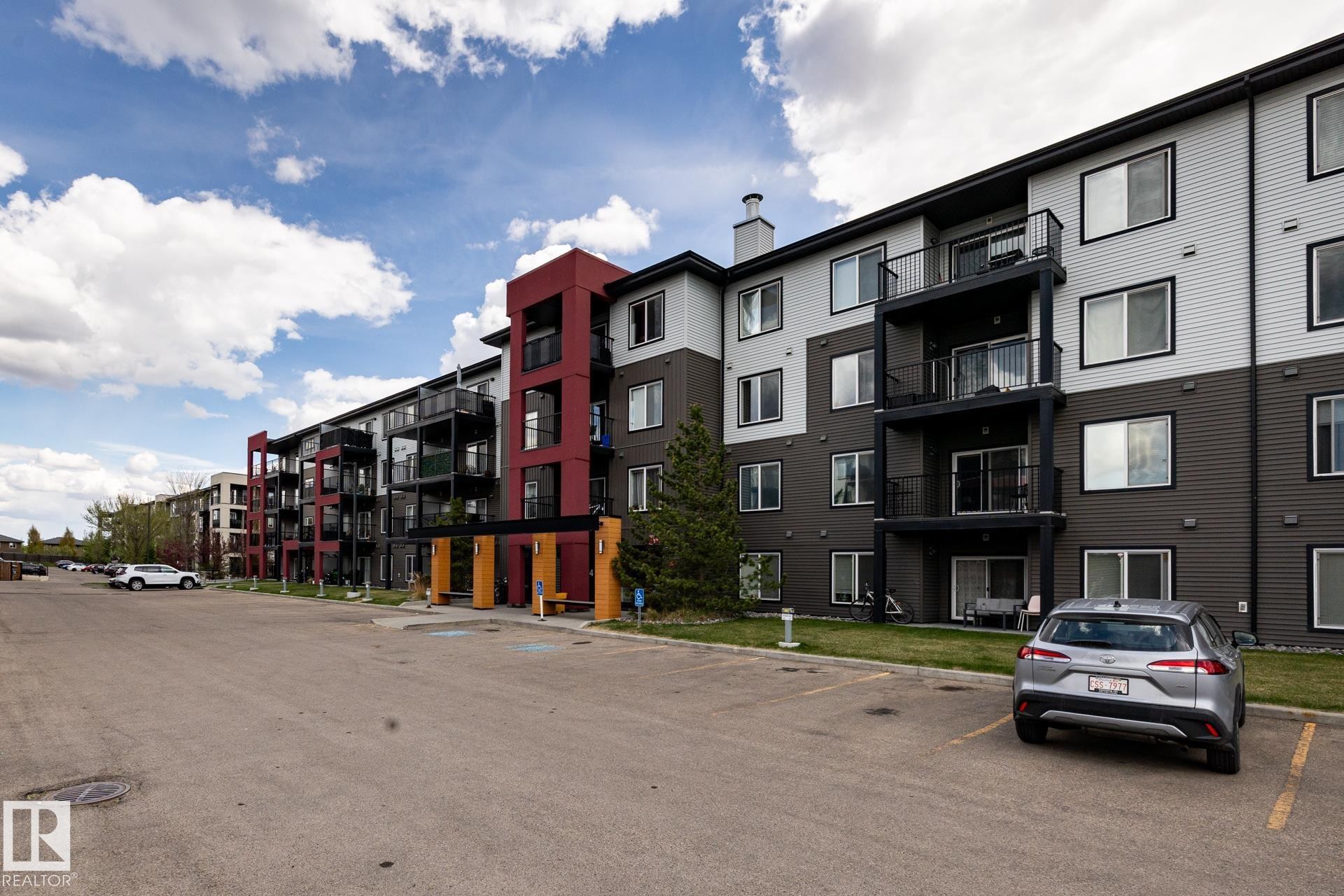 Contemporary multi-story building featuring a facade of light and dark horizontal siding, red architectural accents, multiple private balconies, and a paved parking area - 131 344 Windermere Road, Edmonton, AB - Outdoor With Facade