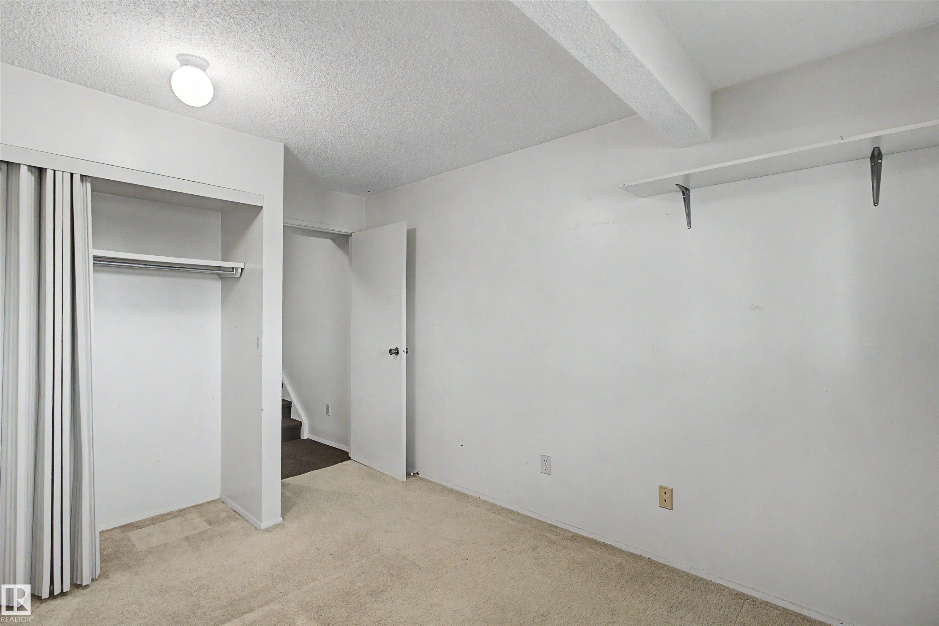 Carpeted room featuring a built-in closet with bi-fold doors, a single ceiling light fixture, exposed ceiling beams, and a wall-mounted shelf with brackets - 5953 40 Avenue, Edmonton, AB