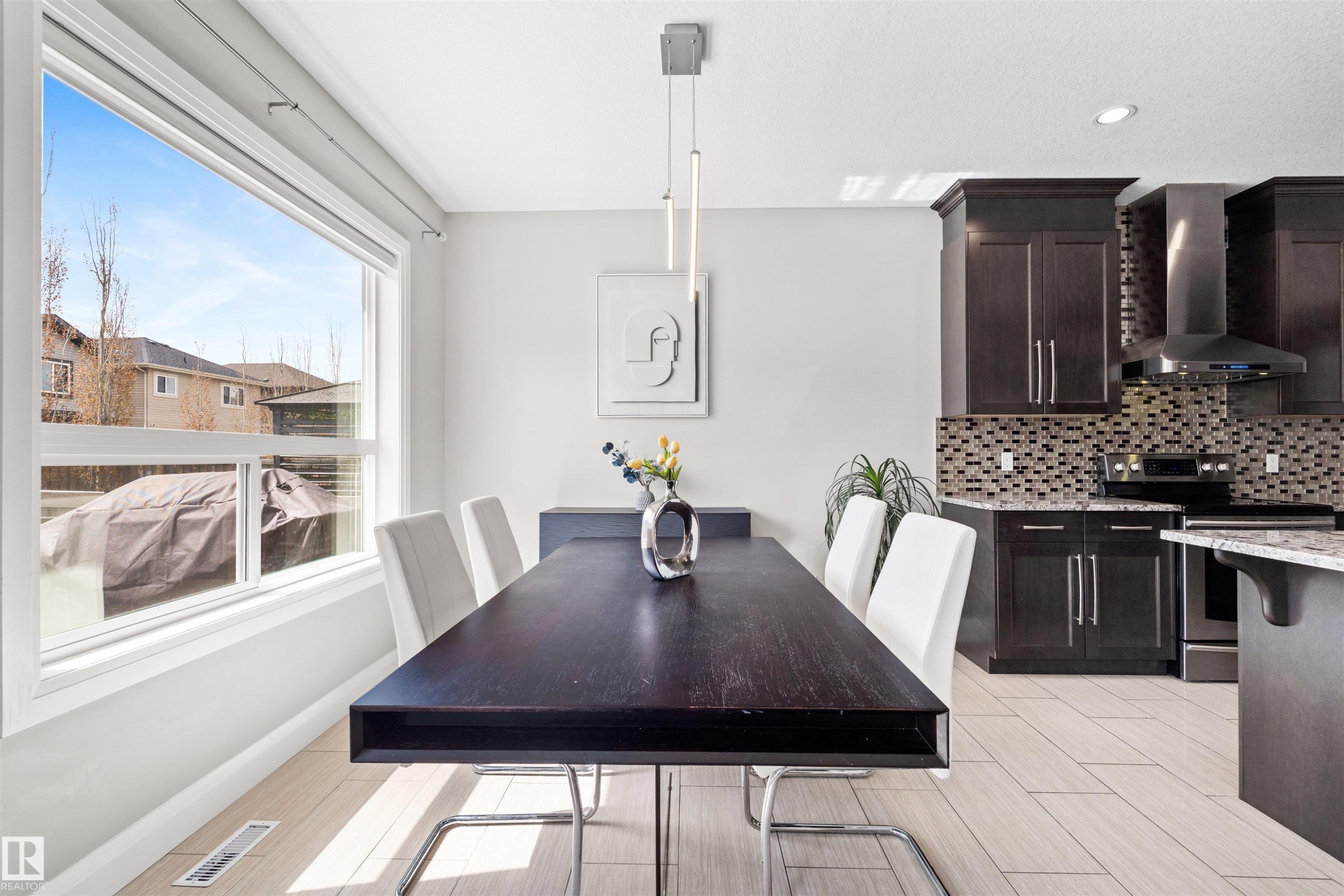 Open-concept dining area featuring a large window, contemporary linear pendant lighting, light wood-finish flooring, and dark cabinetry in the adjacent kitchen - 3807 Claxton Close, Edmonton, AB - Indoor