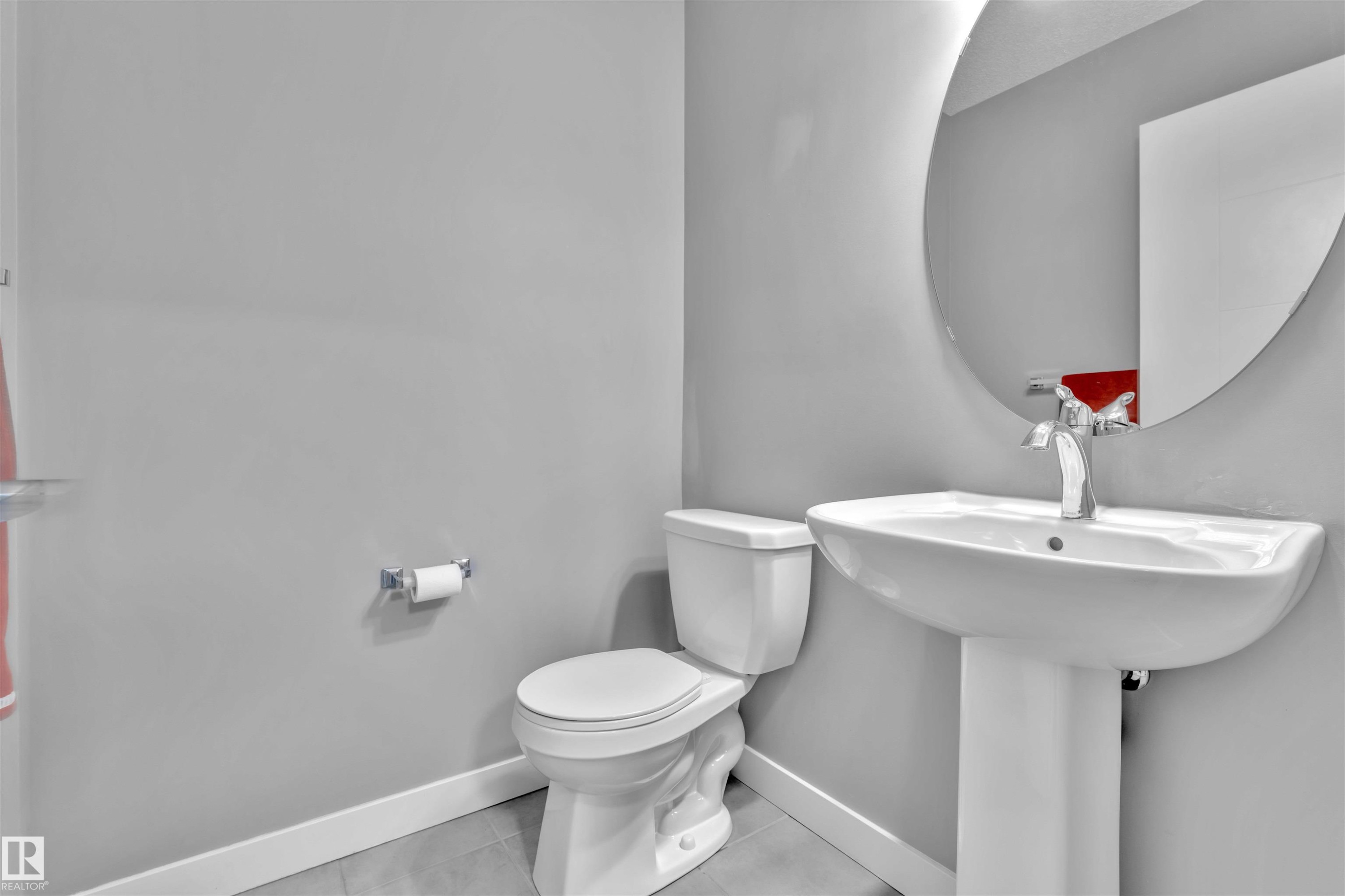 The bathroom features a white pedestal sink with a chrome faucet, a white toilet, and a large round mirror - 8108 217 Street, Edmonton, AB - Indoor Photo Showing Bathroom