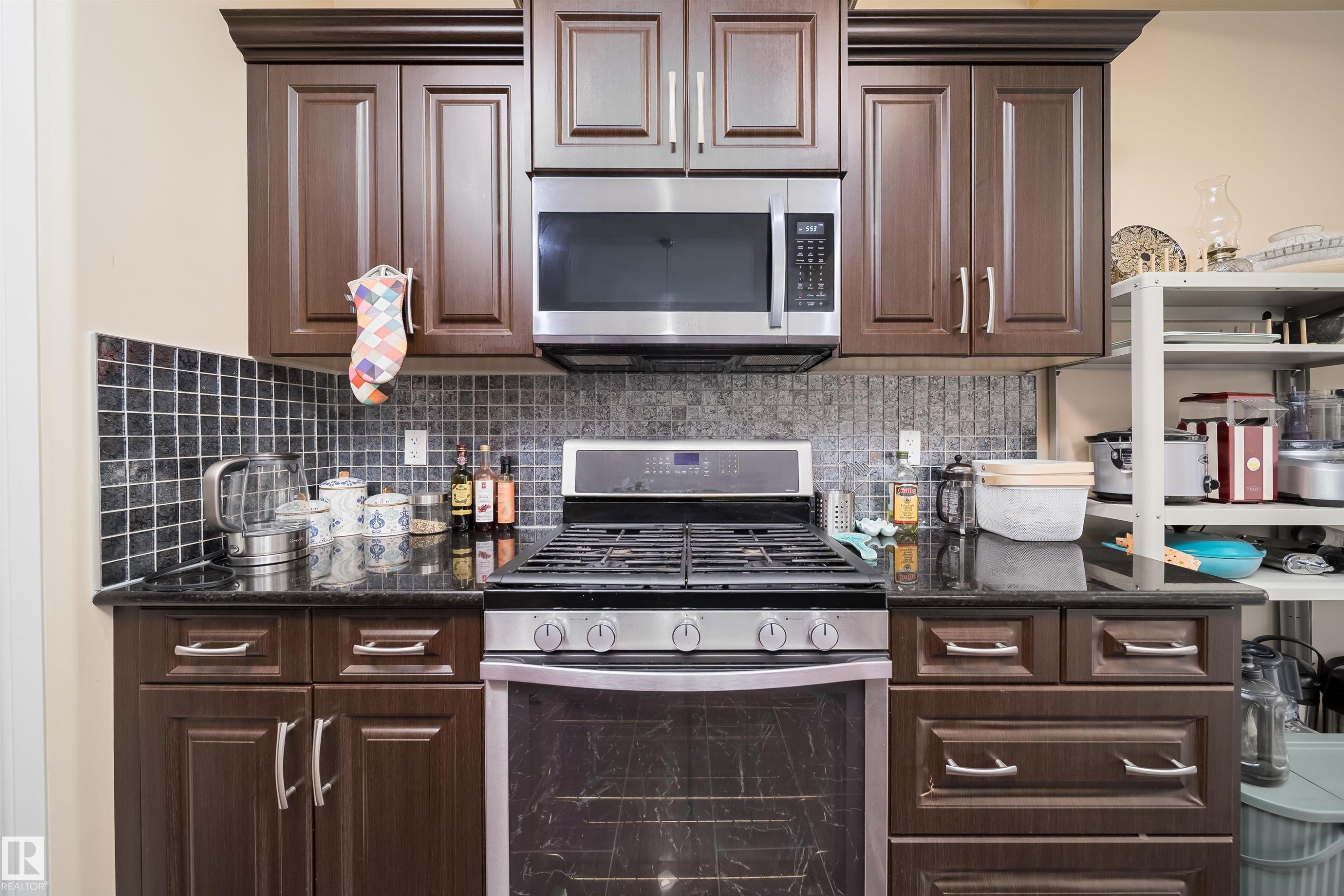 Kitchen featuring dark wood-finish cabinetry, stainless steel oven with gas range, stainless steel microwave, black countertops, and a mosaic tile backsplash - 1508 Cunningham Cape Cape, Edmonton, AB - Indoor Photo Showing Kitchen