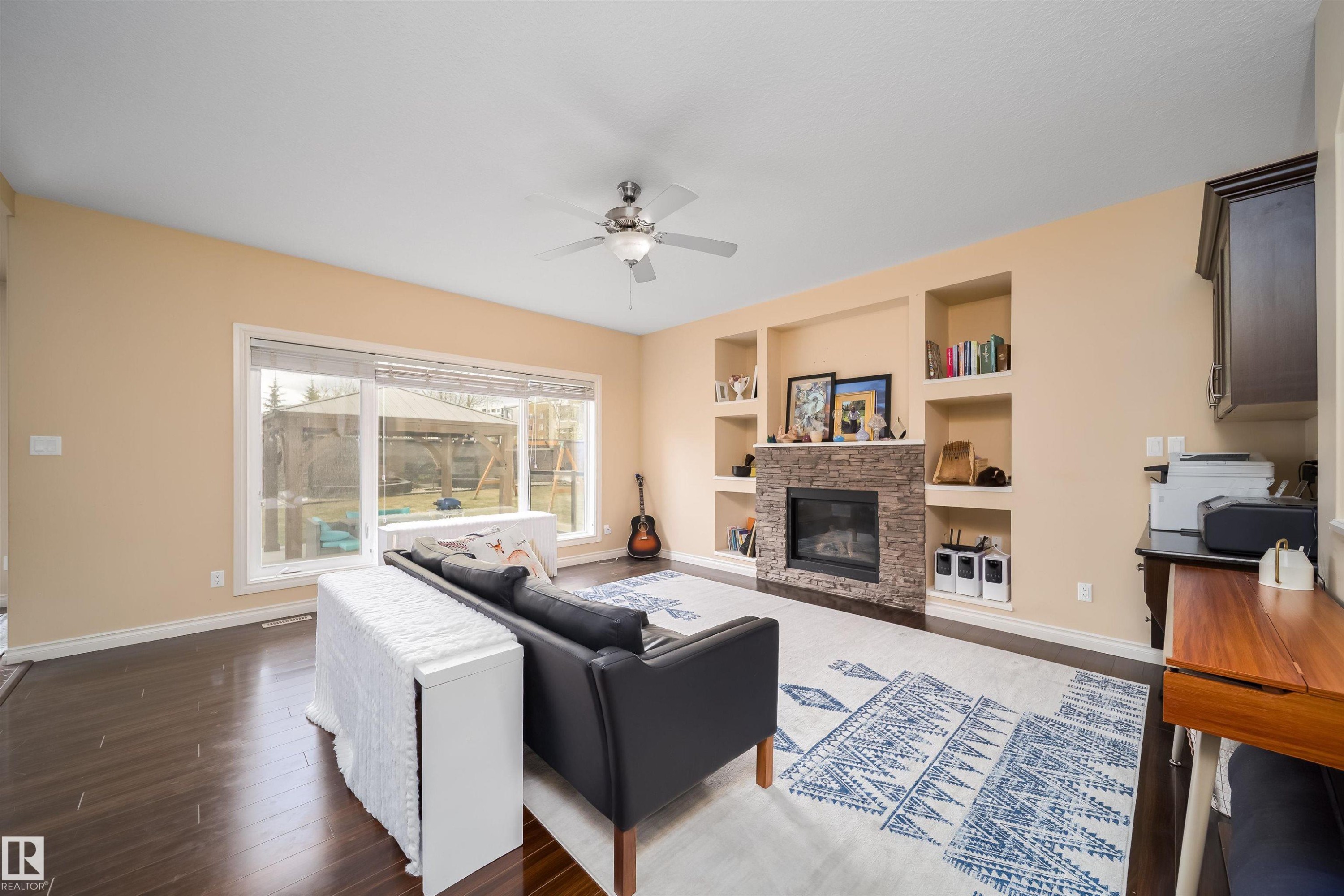 Living area featuring wood-finish flooring, a stone-clad fireplace, integrated shelving, a ceiling fan, and large windows - 1508 Cunningham Cape Cape, Edmonton, AB - Indoor Photo Showing Living Room With Fireplace