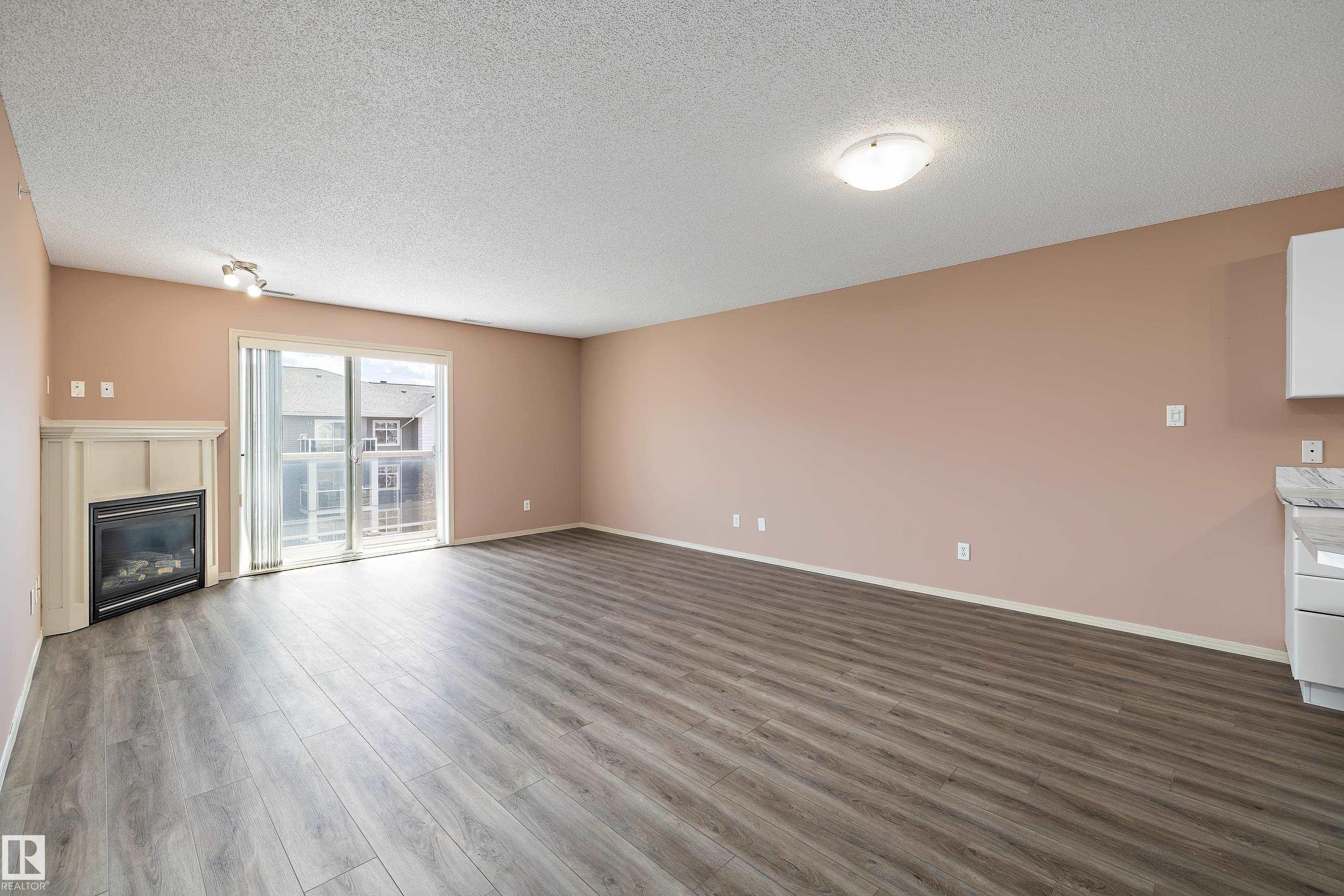 Spacious room featuring wood-finish flooring, a built-in electric fireplace with a white mantel, and a sliding glass door providing access to an outdoor area - 406 16035 132 Street, Edmonton, AB - Indoor With Fireplace
