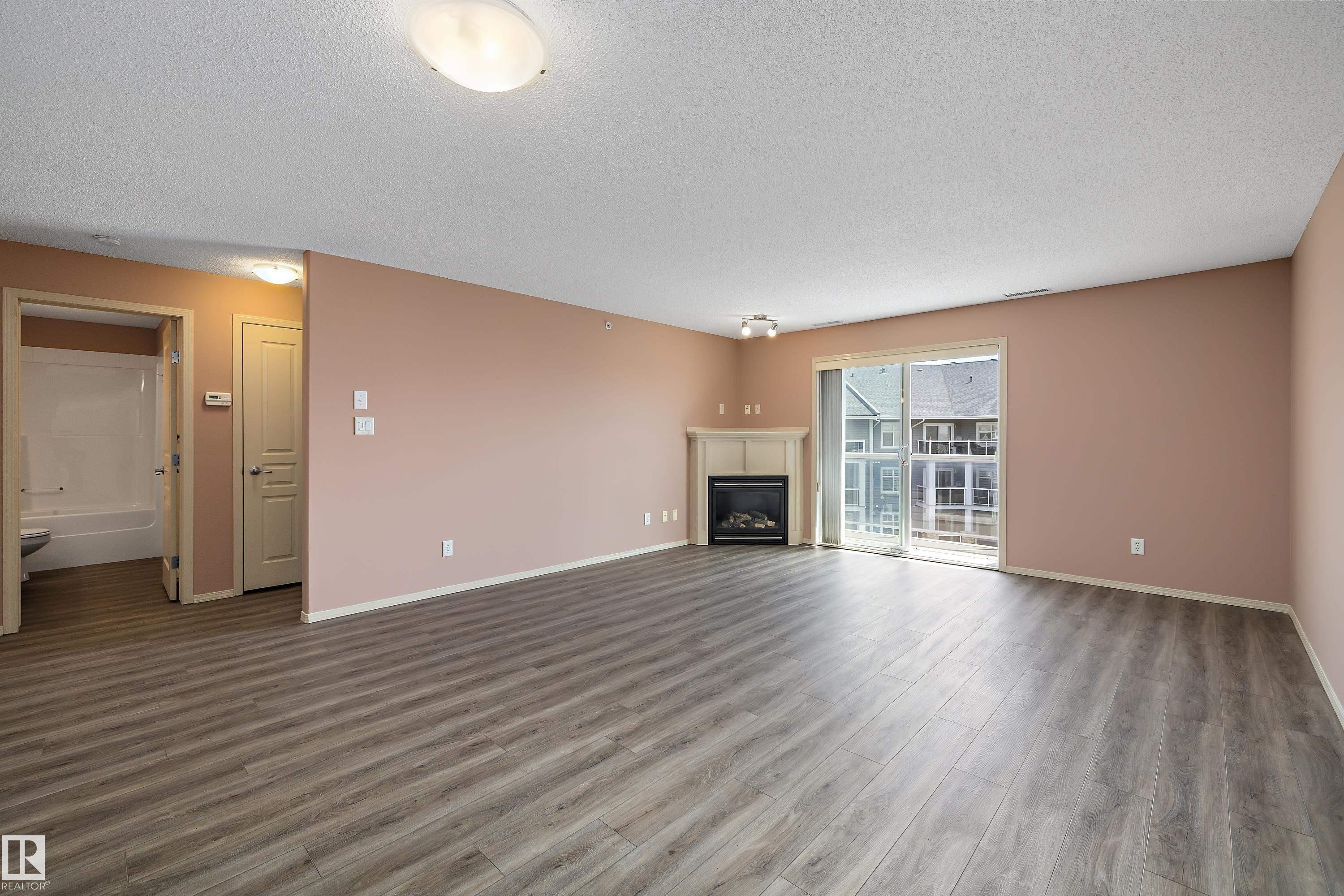 Spacious living area featuring wood-finish flooring, a corner fireplace with a white mantel, and sliding glass doors opening to the exterior - 406 16035 132 Street, Edmonton, AB - Indoor Photo Showing Living Room With Fireplace