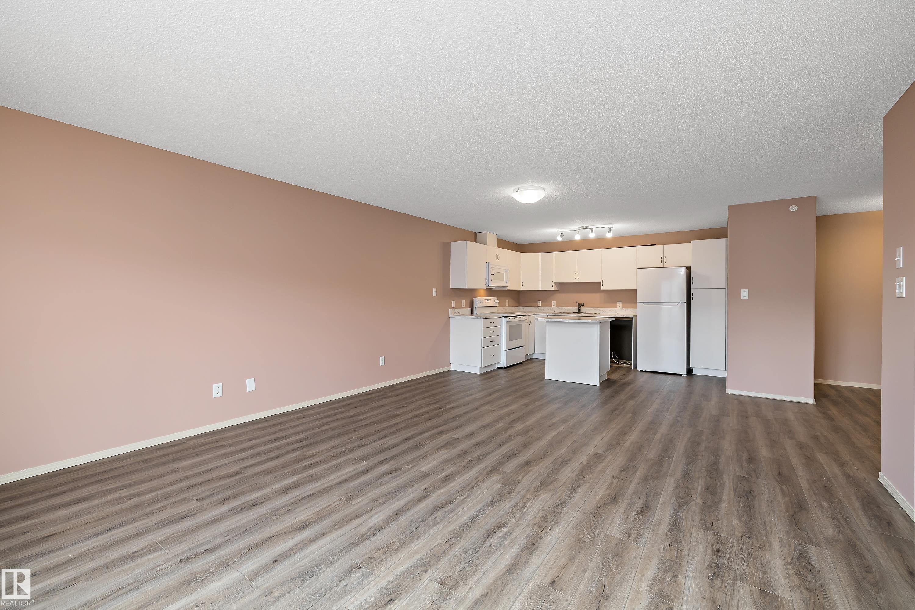 Open-concept living space featuring wood-finish flooring, a contemporary kitchen with white cabinetry and appliances, a central kitchen island, and recessed lighting - 406 16035 132 Street, Edmonton, AB - Indoor Photo Showing Kitchen