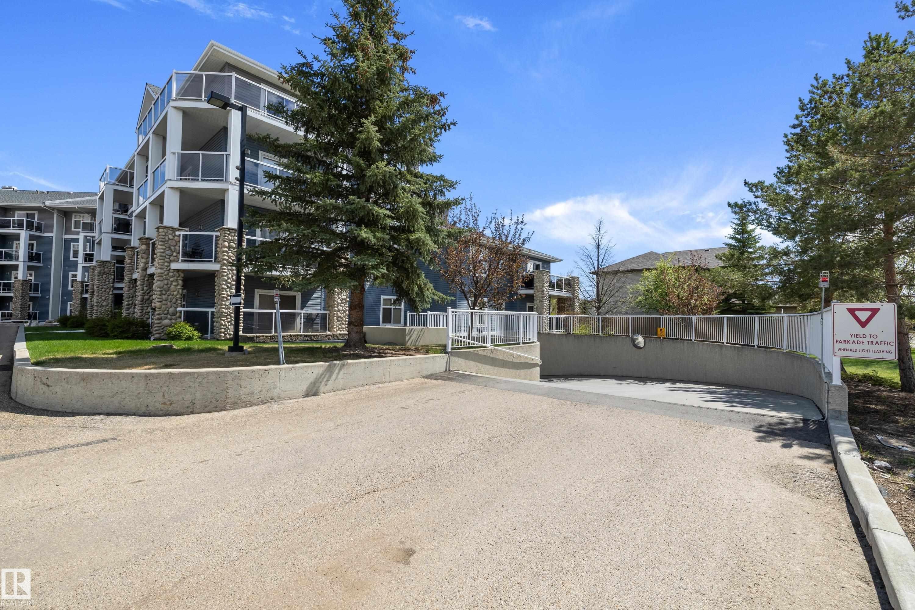 Multi-story building featuring stacked balconies with glass railings, stone veneer accents, and a sloped concrete entrance to subterranean parking - 406 16035 132 Street, Edmonton, AB - Outdoor With Balcony