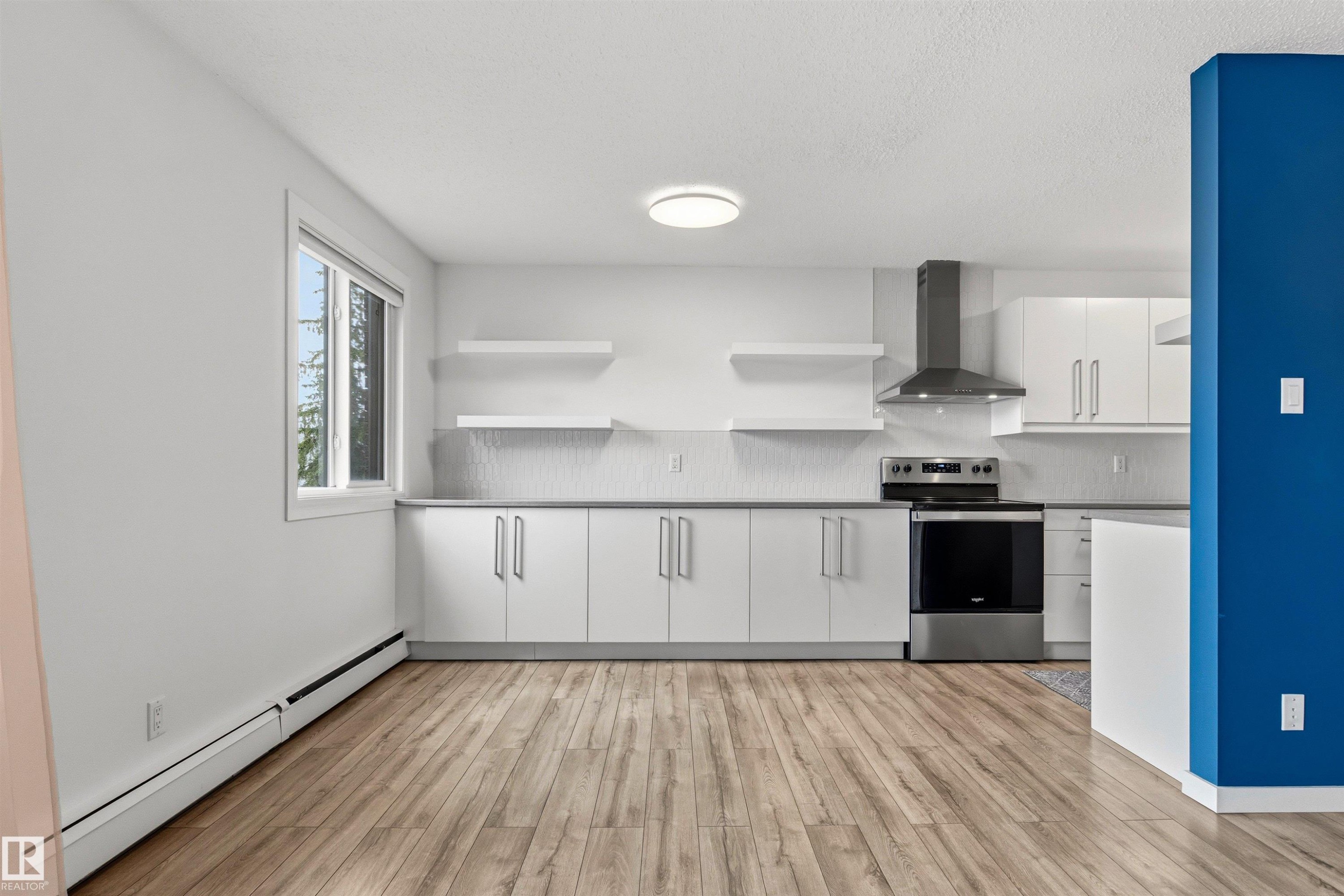 Modern kitchen featuring light wood-finish flooring, white cabinetry with silver hardware, a stainless steel range with overhead hood, a window, and open shelving - 217 14819 51 Avenue, Edmonton, AB