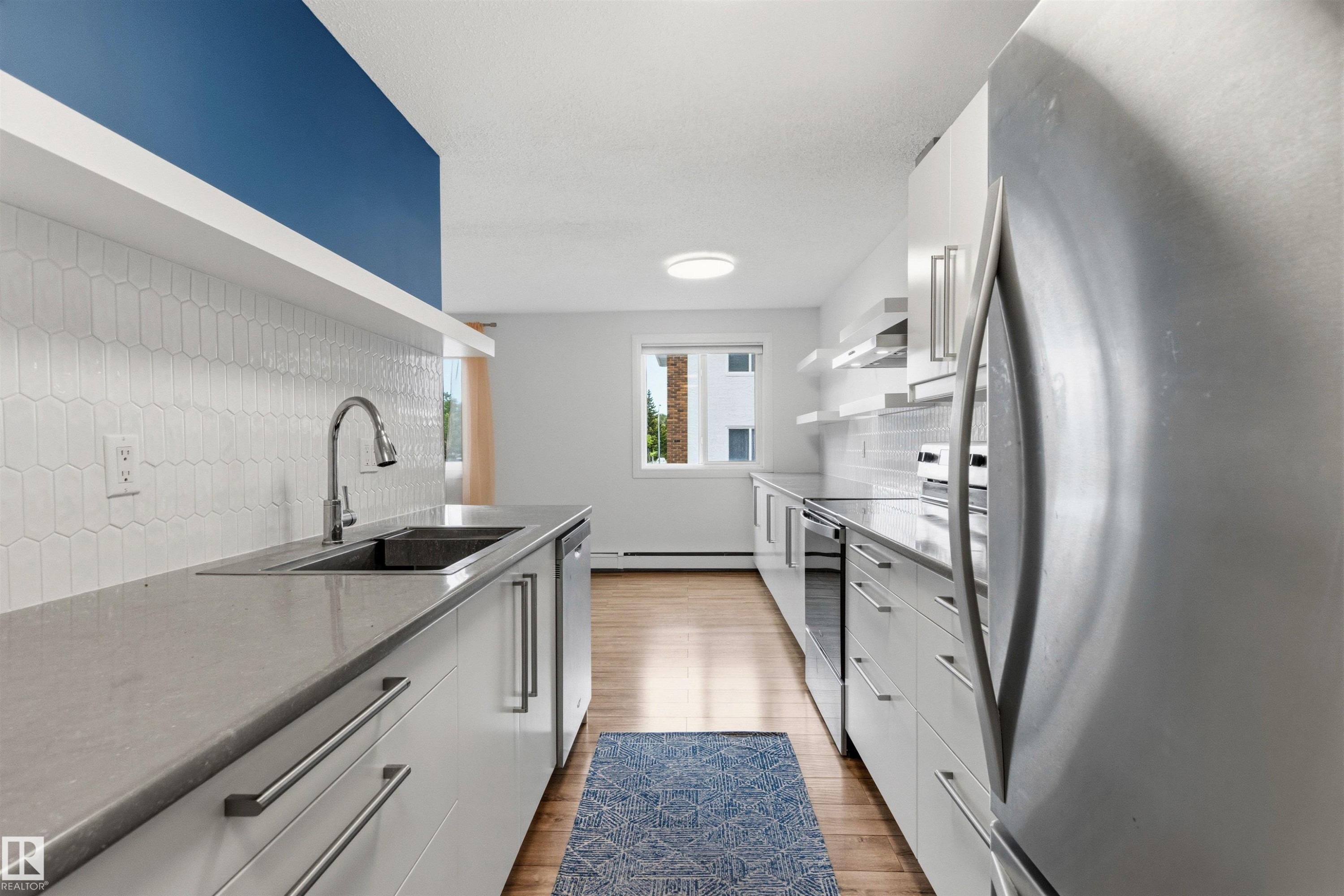 Galley kitchen featuring white cabinetry, light-toned countertops, a stainless steel sink with a gooseneck faucet, a white picket tile backsplash, and a full suite of stainless steel appliances - 217 14819 51 Avenue, Edmonton, AB