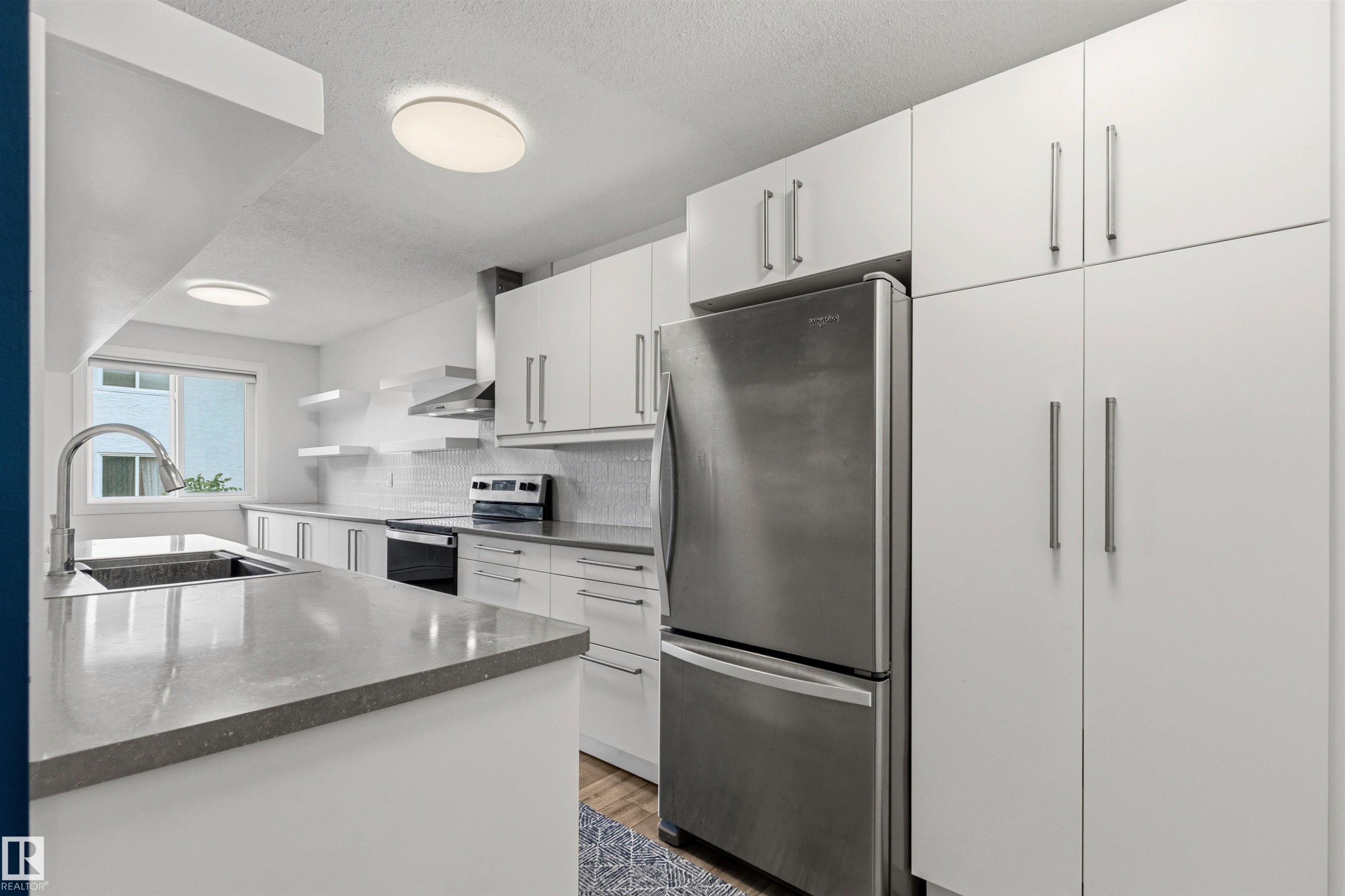 Modern kitchen featuring extensive white cabinetry, a stainless steel refrigerator, and a kitchen island with an undermount sink - 217 14819 51 Avenue, Edmonton, AB