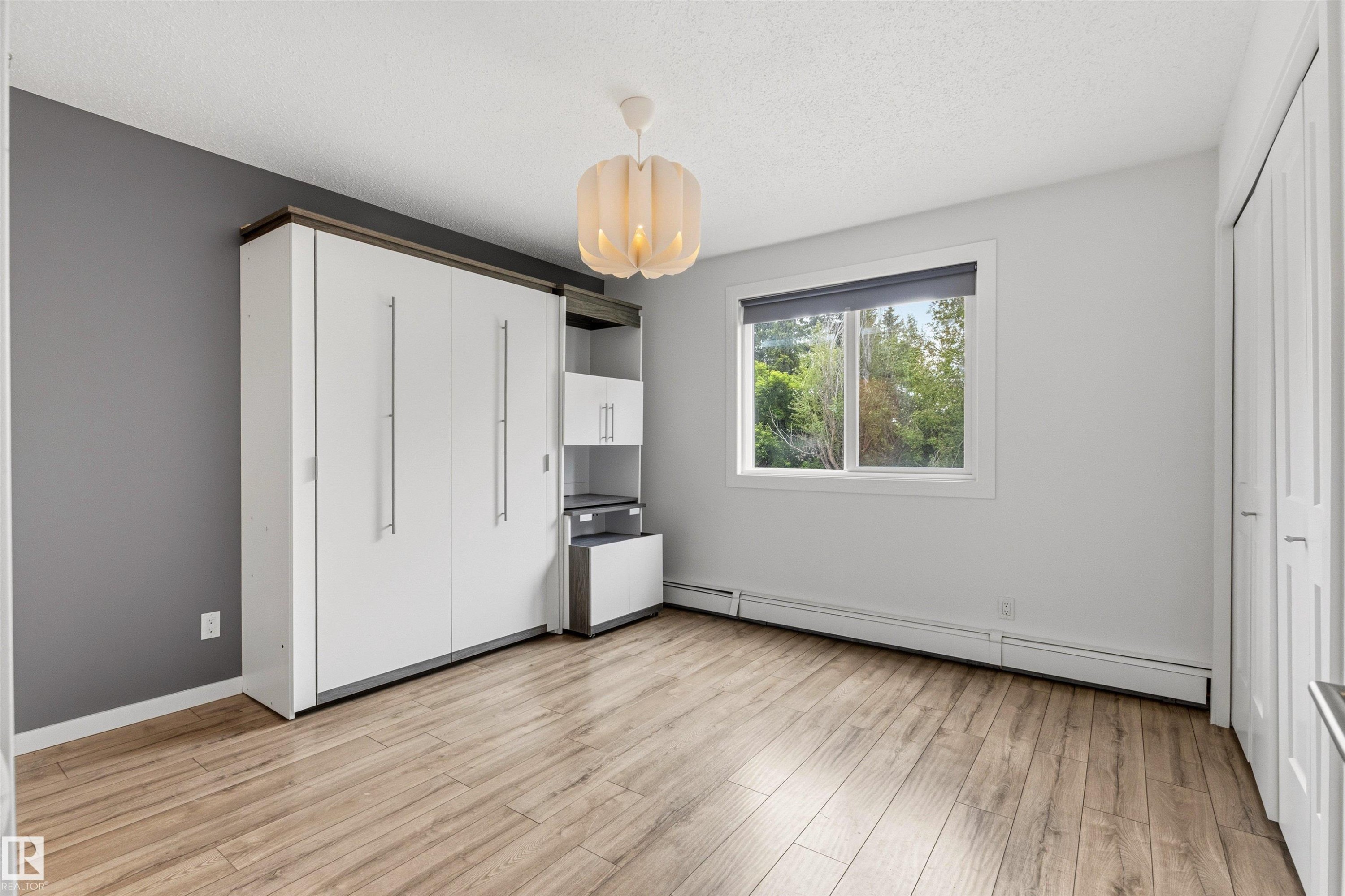 Room featuring wood-finish flooring, a built-in white cabinetry system with integrated shelving, and a prominent window with roller blinds - 217 14819 51 Avenue, Edmonton, AB
