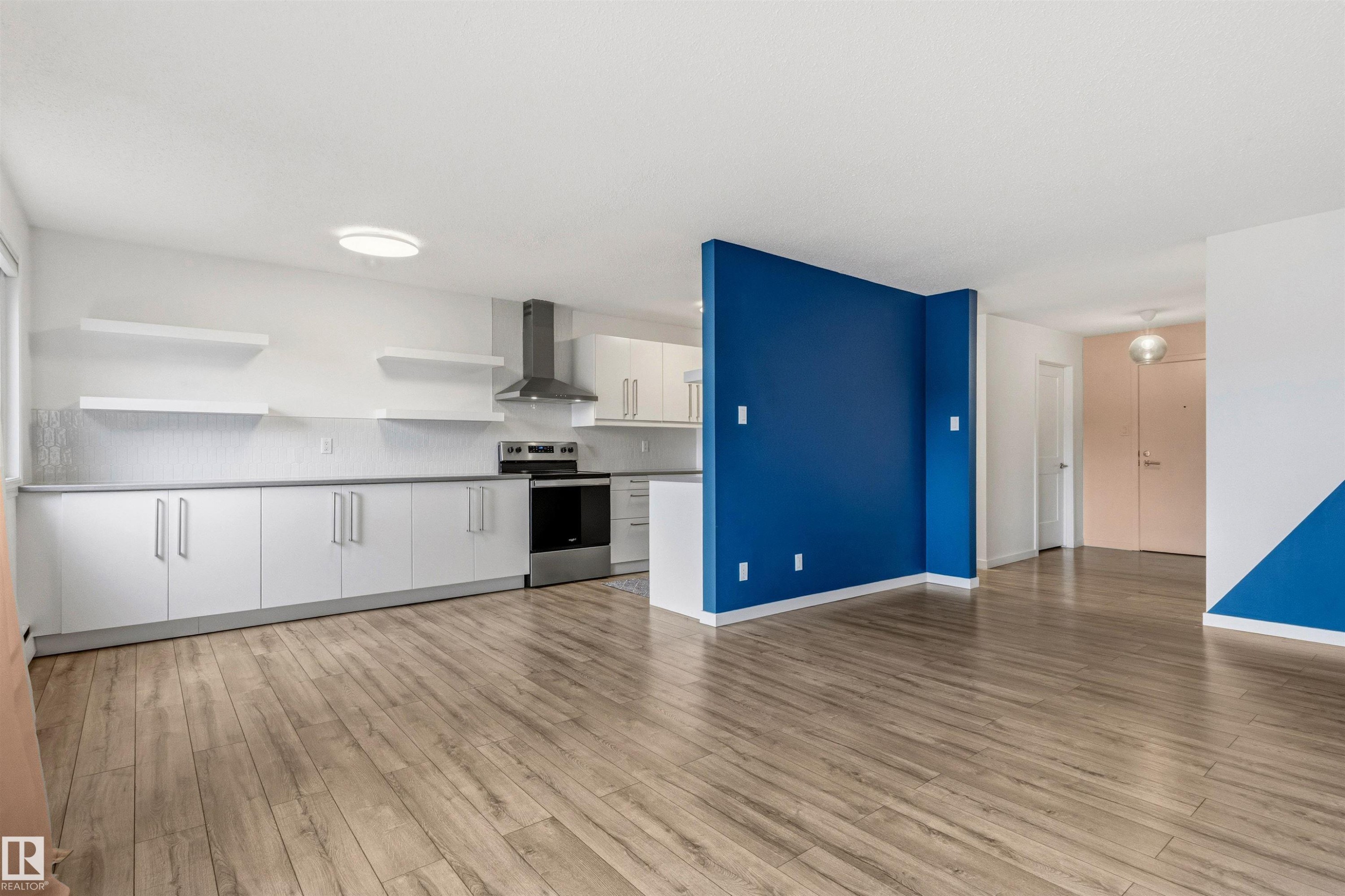 Open-concept living space featuring wood-finish flooring, a kitchen with white cabinetry and stainless steel appliances, and a distinctive blue accent wall - 217 14819 51 Avenue, Edmonton, AB