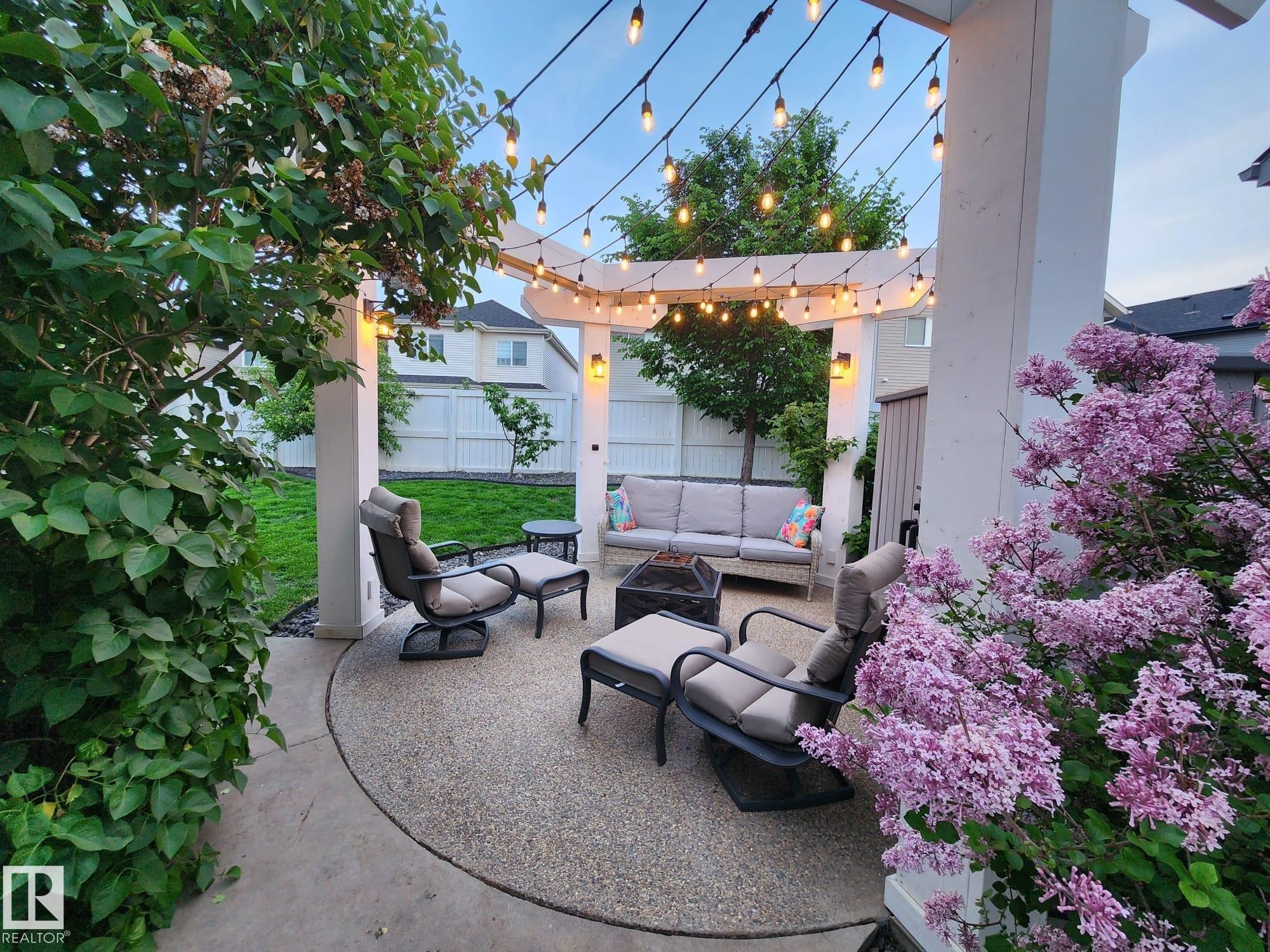 Patio area featuring an exposed aggregate concrete surface, a white pergola with string lighting, exterior wall sconces, and mature landscaping - 22933 94A Avenue, Edmonton, AB - Outdoor With Deck Patio Veranda With Exterior