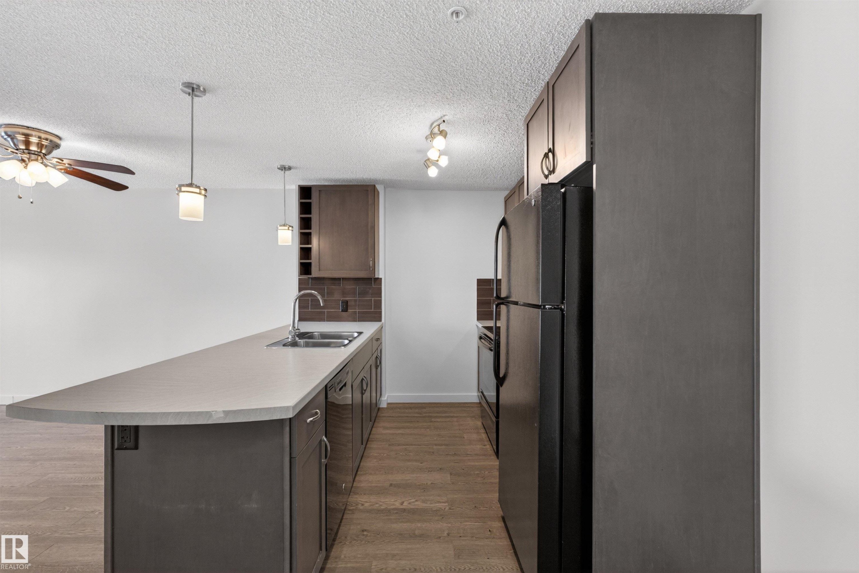 Kitchen featuring a spacious island with an integrated sink, dark wood-finish cabinetry, a subway tile backsplash, and black appliances - 215 6084 Stanton Drive, Edmonton, AB - Indoor Photo Showing Kitchen With Double Sink