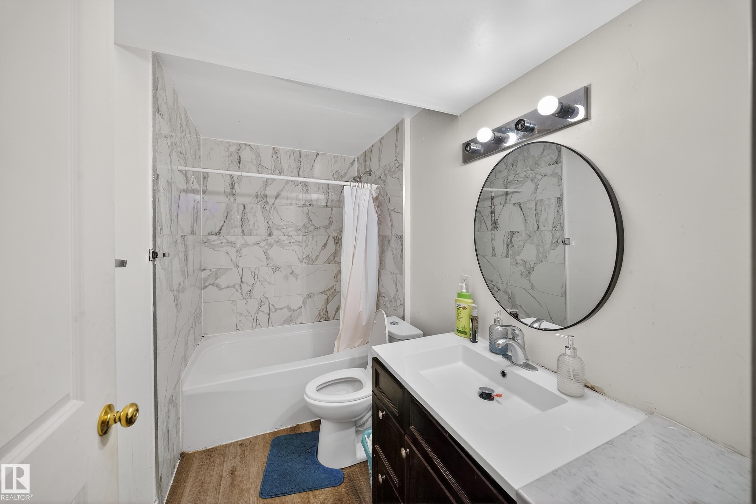This bathroom features a full-sized bathtub with white and grey marble-style tiled walls, a dark wood vanity with a white countertop and integrated sink, and a circular mirror with a modern light fixture overhead - 3517 25 Street, Edmonton, AB - Indoor Photo Showing Bathroom
