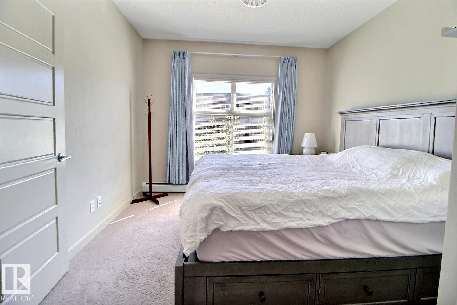 Neutral-toned room with light-colored carpeting and a window featuring light blue drapes - 327 508 Griesbach Parade, Edmonton, AB - Indoor Photo Showing Bedroom