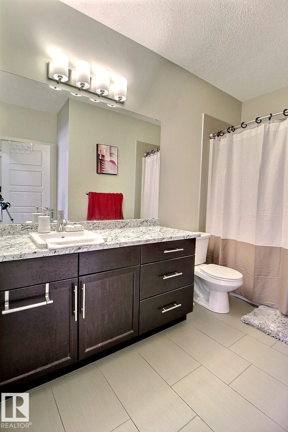 Bathroom featuring a dark wood-finish vanity with ample storage, a light-toned countertop, a full-width mirror, and a modern four-light fixture - 327 508 Griesbach Parade, Edmonton, AB - Indoor Photo Showing Bathroom