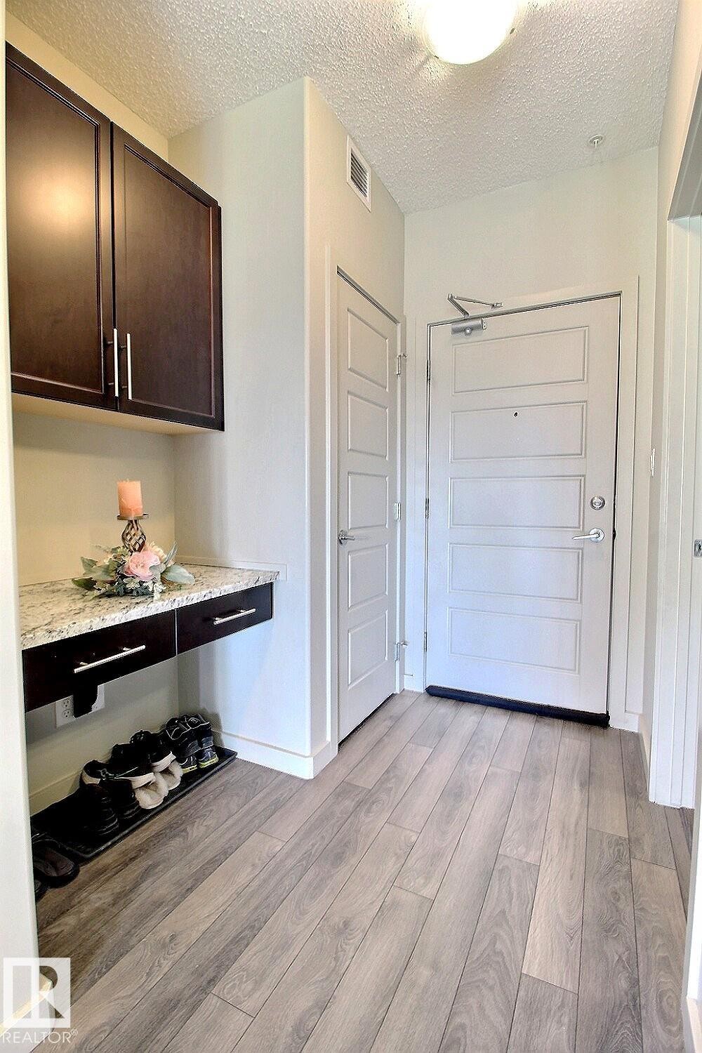 Entryway featuring wood-finish flooring, a wall-mounted dark wood-finish cabinet with a granite-look countertop, recessed lighting, and a solid entry door with a multi-paneled design - 327 508 Griesbach Parade, Edmonton, AB - Indoor Photo Showing Other Room