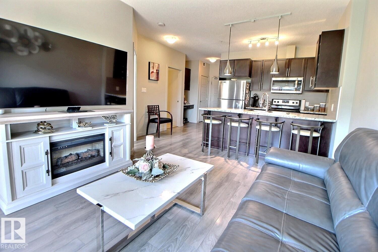 Open-concept living area featuring wood-finish flooring, a kitchen with dark cabinetry, stainless steel appliances, and a breakfast bar with pendant lighting - 327 508 Griesbach Parade, Edmonton, AB - Indoor