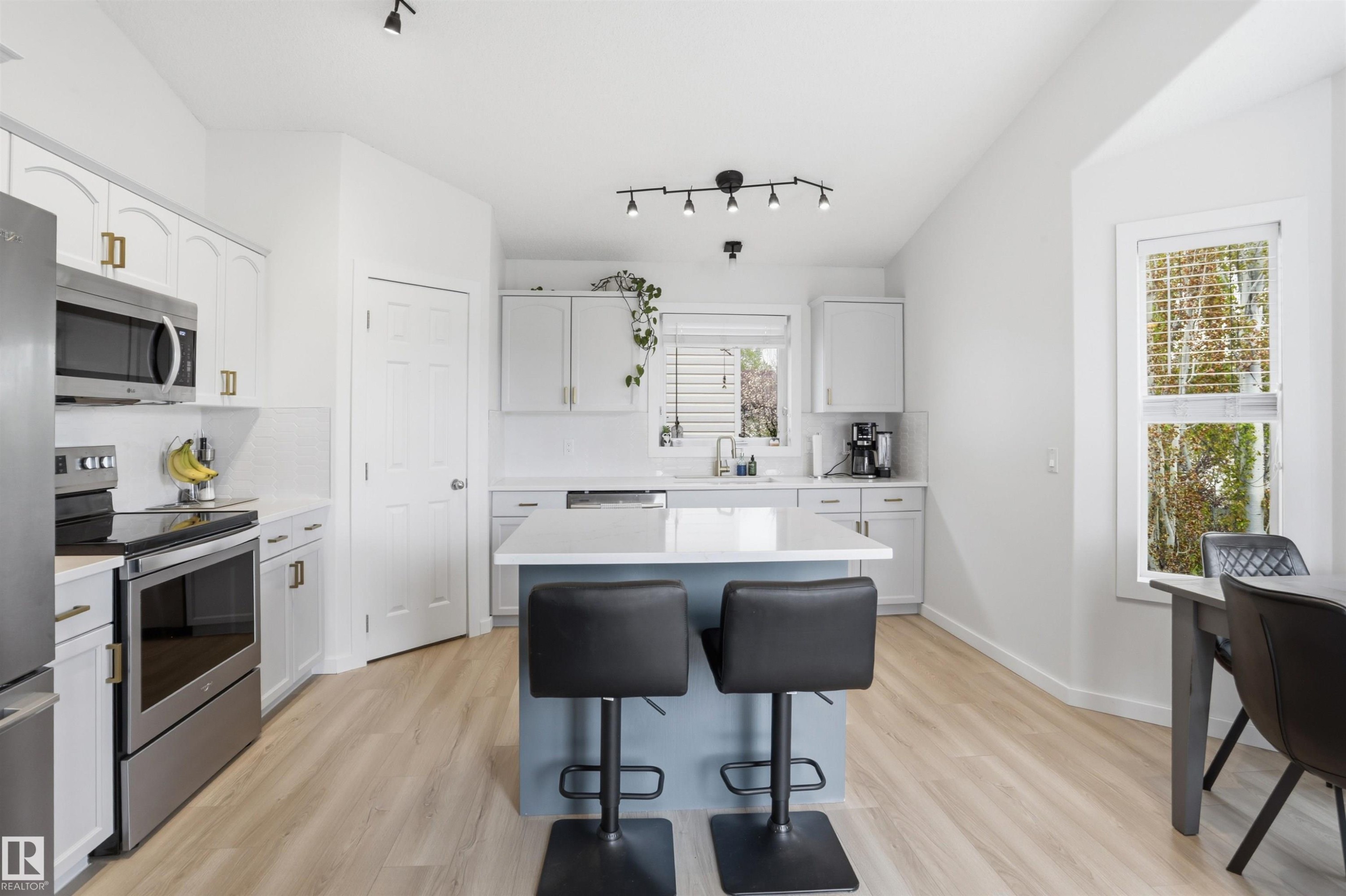 Bright kitchen featuring a central island, stainless steel appliances, light-toned wood-finish flooring, white cabinetry with gold-finish hardware, and track lighting - 396 Kananaskis Way, Devon, AB - Indoor Photo Showing Kitchen