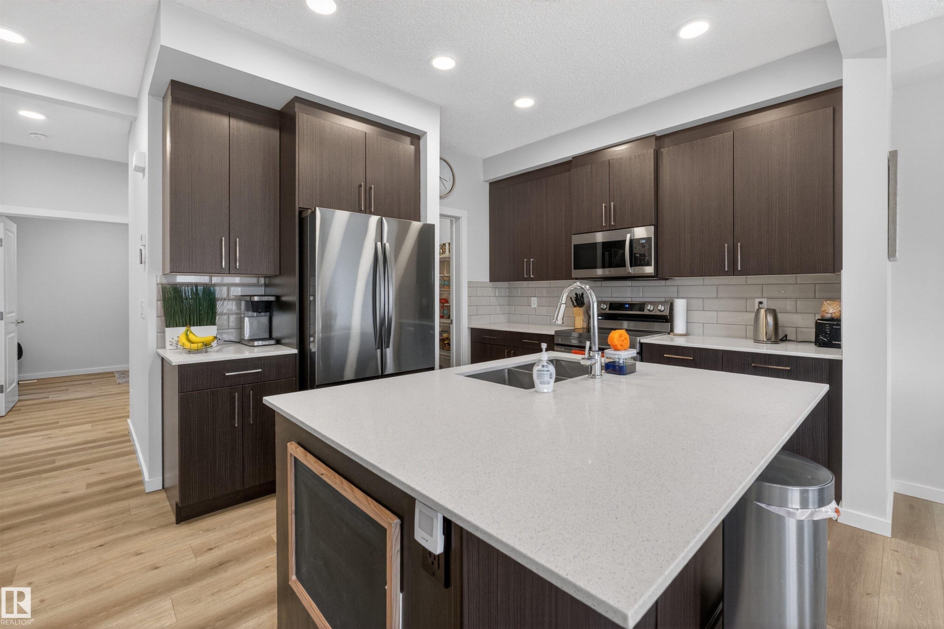Kitchen featuring an expansive island with a light-toned countertop, dark wood-finish cabinetry, stainless steel appliances, a subway tile backsplash, and recessed lighting - 332 Juniper Cove, Leduc, AB - Indoor Photo Showing Kitchen With Upgraded Kitchen