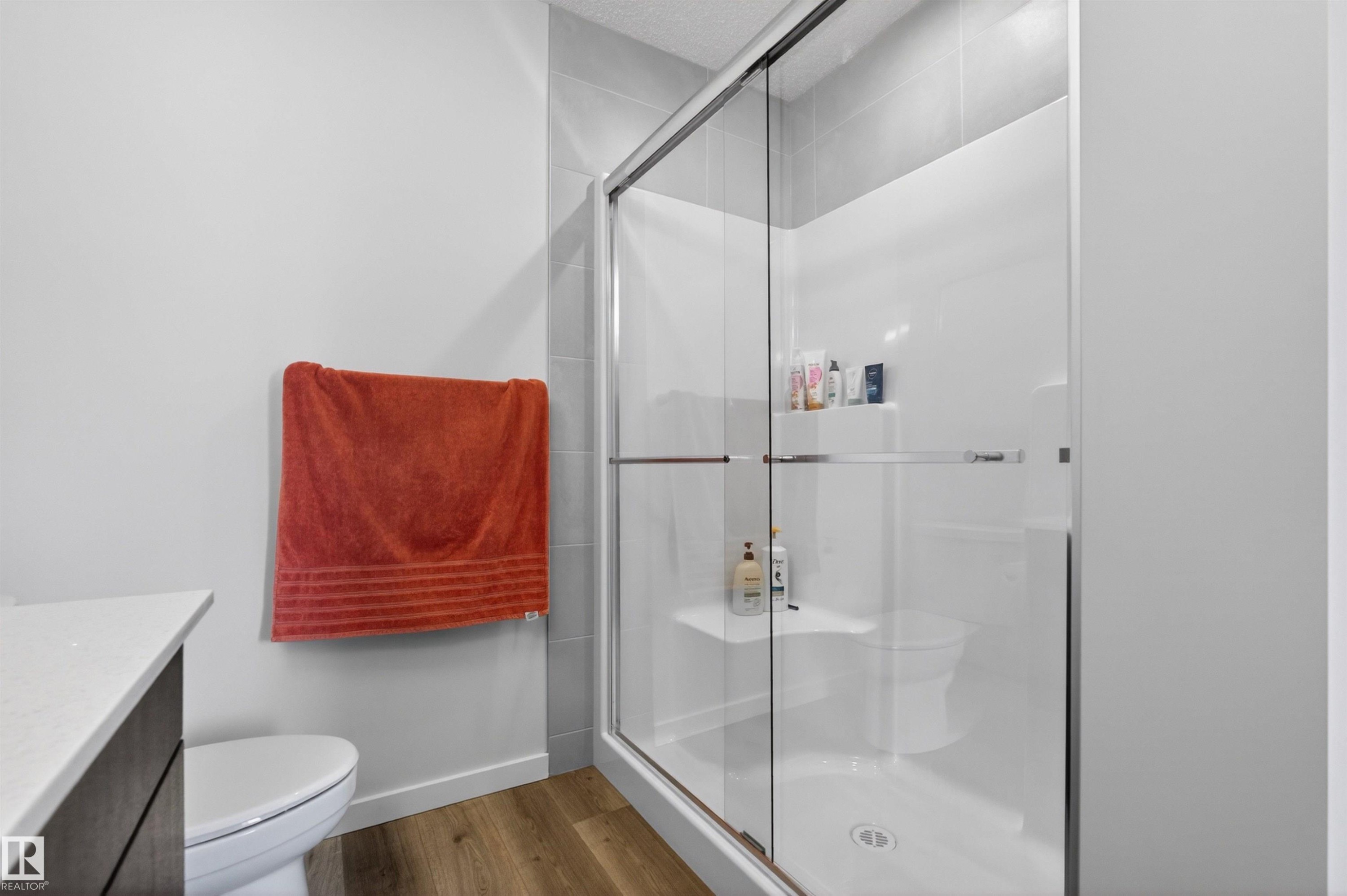 Framed glass shower enclosure with a built-in shower bench and shelf, complemented by a white toilet and wood-finish flooring - 332 Juniper Cove, Leduc, AB - Indoor Photo Showing Bathroom