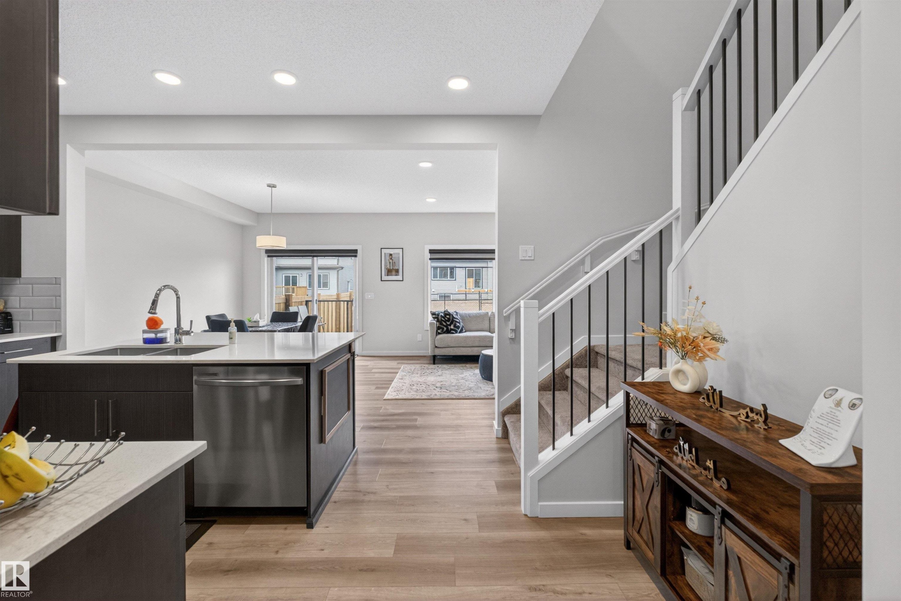 Open-concept living area featuring wood-finish flooring, recessed lighting, and a contemporary kitchen island with a stainless steel dishwasher and sink - 332 Juniper Cove, Leduc, AB - Indoor Photo Showing Kitchen With Upgraded Kitchen