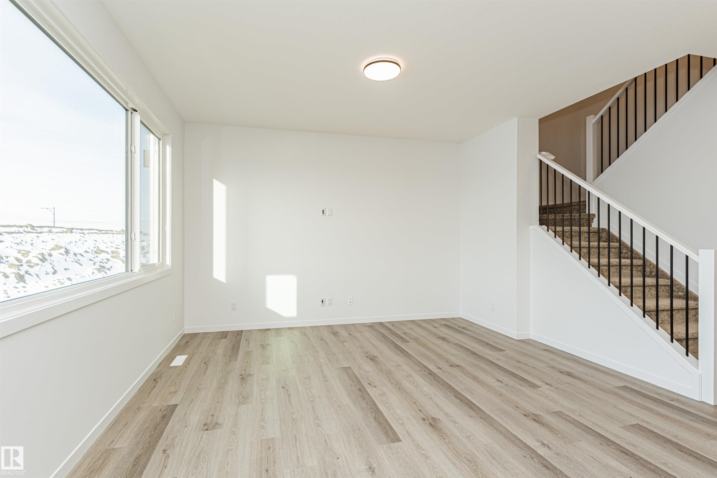 Spacious room featuring light-colored flooring, a large window, and a staircase with dark balusters and white risers - 2355 Egret Way, Edmonton, AB - Indoor Photo Showing Other Room