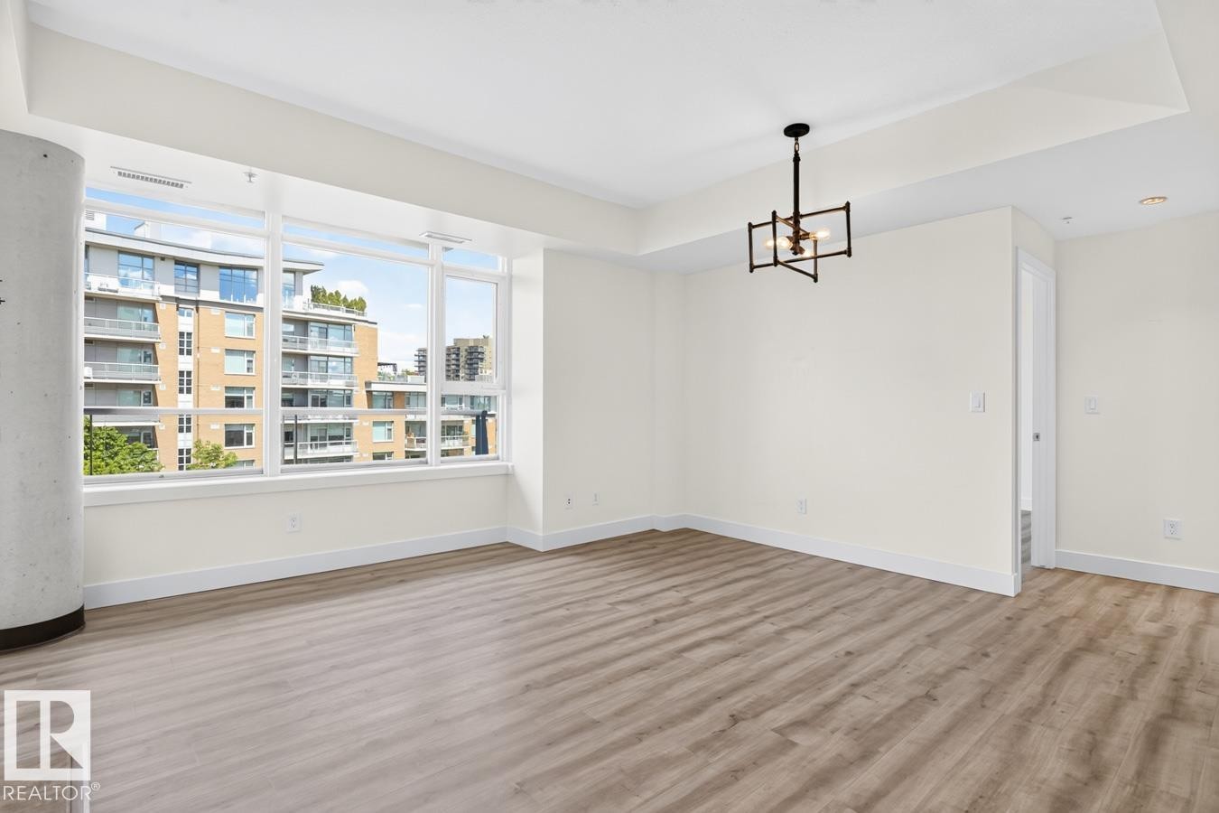 Spacious living area featuring expansive windows, wood-finish flooring, a recessed tray ceiling, white baseboards, and a modern geometric chandelier - #407 2504 109 St Nw, Edmonton, AB - Indoor Photo Showing Other Room