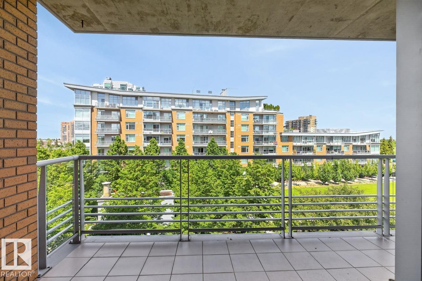 Balcony featuring tile flooring and metal railings, overlooking a tree-lined green space with urban buildings in the background - #407 2504 109 St Nw, Edmonton, AB - Outdoor With Balcony