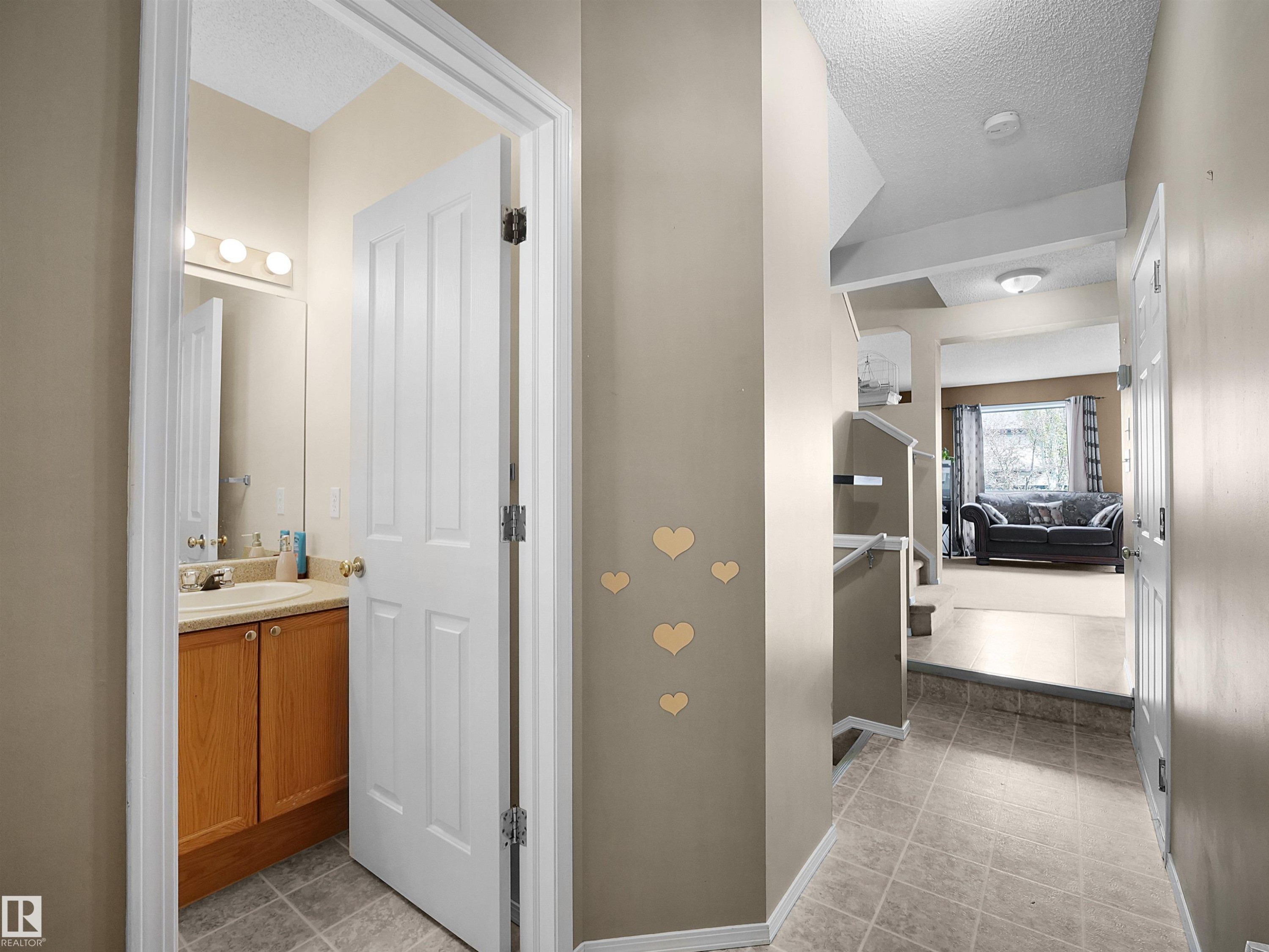 Hallway featuring neutral wall paint, tiled flooring, an open doorway revealing a bathroom with a wood-finish vanity, and a partial view into a carpeted living space - 16121 131A Street, Edmonton, AB - Indoor Photo Showing Other Room
