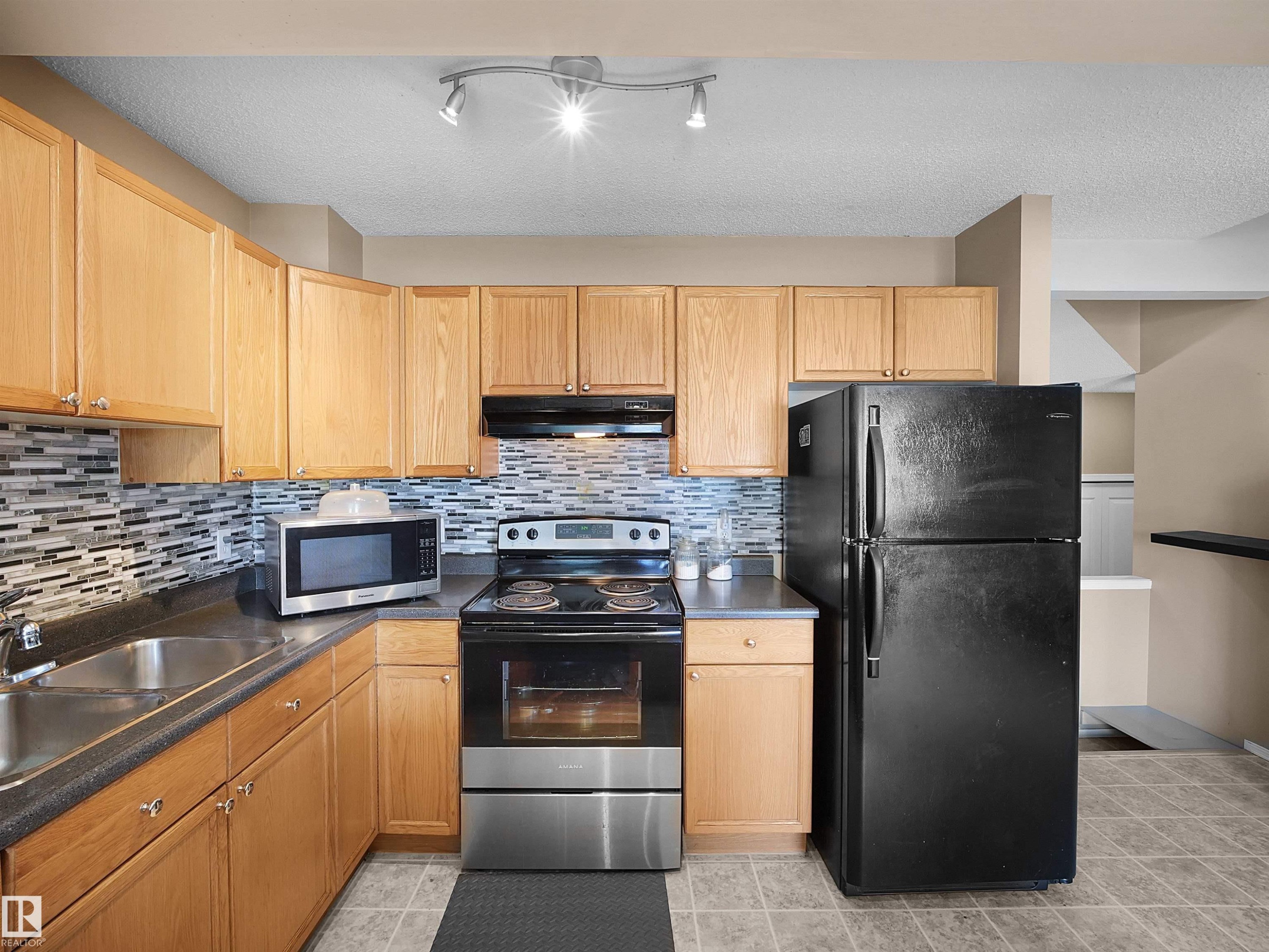 Kitchen featuring light wood cabinetry, dark countertops, a mosaic tile backsplash, stainless steel sink, and track lighting - 16121 131A Street, Edmonton, AB - Indoor Photo Showing Kitchen With Double Sink