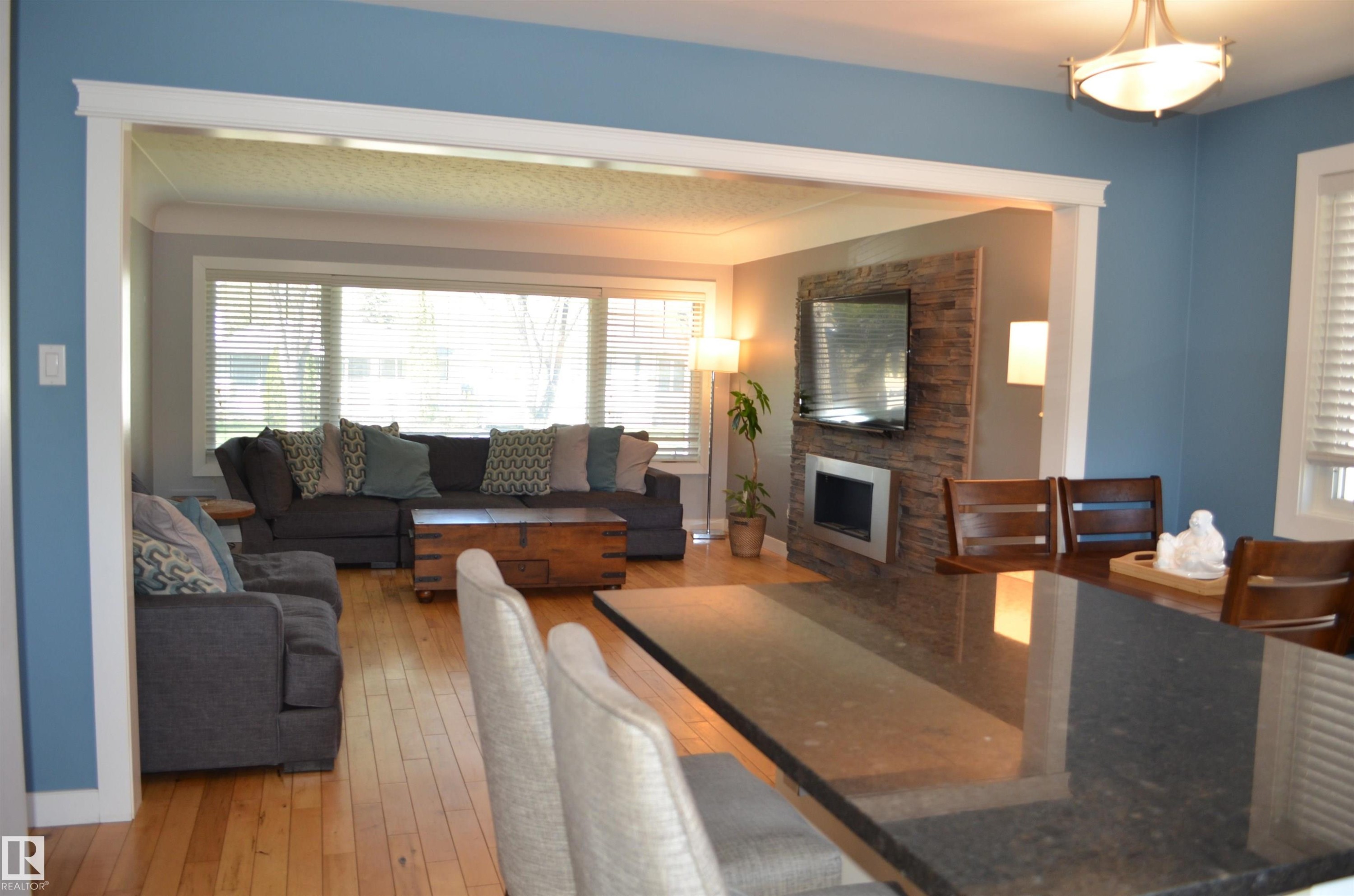 Open-concept living space featuring wood-finish flooring, a contemporary fireplace with a stacked stone-look surround, and a large front window with horizontal blinds - 10527 81 Street, Edmonton, AB - Indoor Photo Showing Living Room With Fireplace