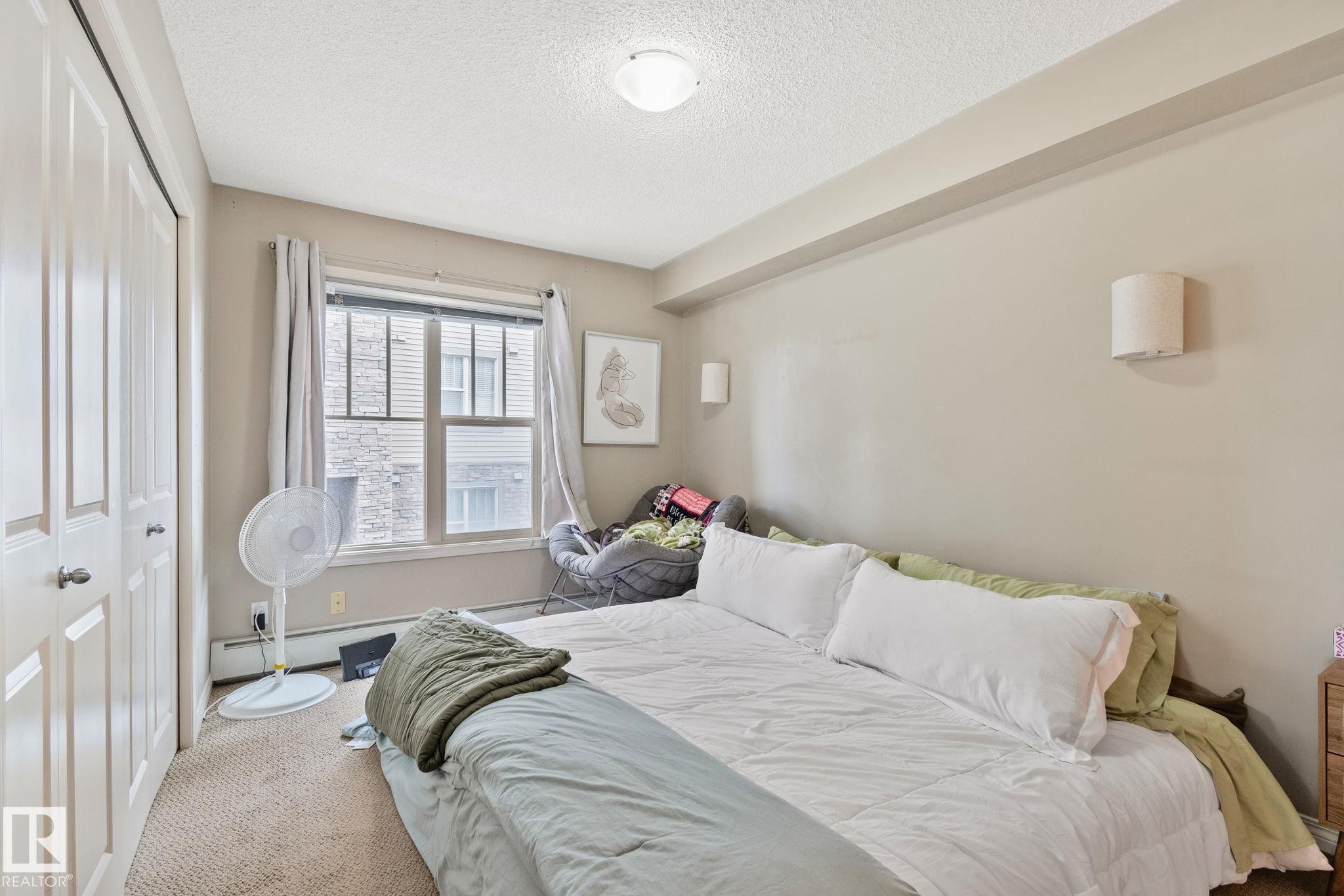 Bedroom featuring a double-door closet with panel detailing, a large window, and wall-mounted sconce lighting - 206 103 Ambleside Drive, Edmonton, AB - Indoor Photo Showing Bedroom