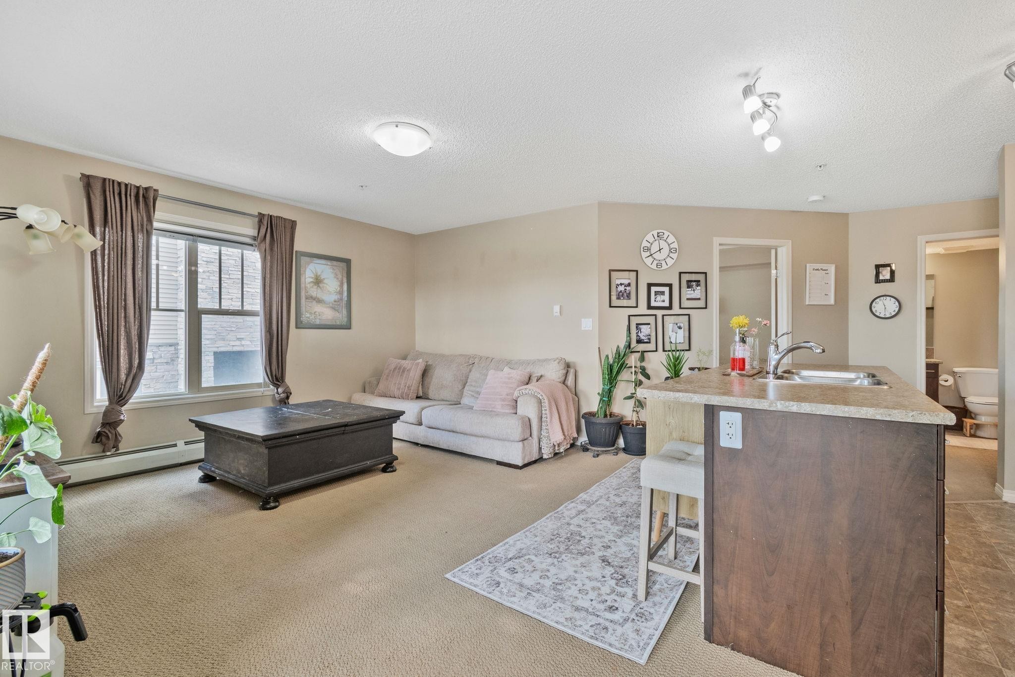 Open-concept living space featuring neutral carpet flooring, a large window, and a kitchen island with a built-in sink and countertop - 206 103 Ambleside Drive, Edmonton, AB - Indoor Photo Showing Living Room