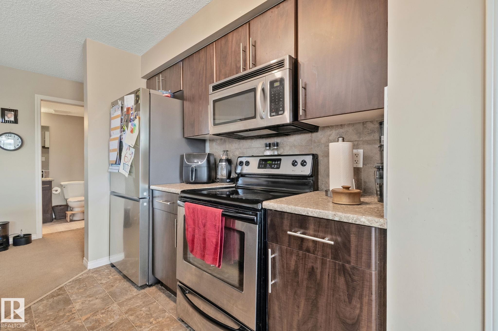 Kitchen featuring wood-finish cabinetry, stainless steel appliances, speckled countertops, and a gray tiled backsplash - 206 103 Ambleside Drive, Edmonton, AB - Indoor Photo Showing Kitchen
