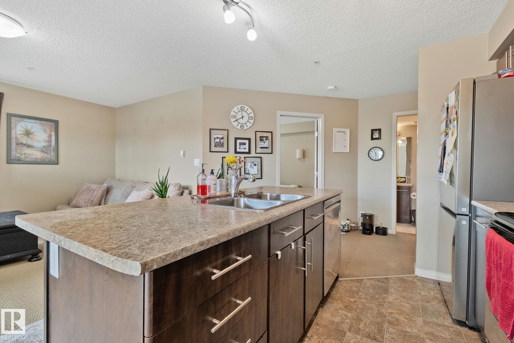Kitchen island featuring a double basin sink, dark wood-finish cabinetry with silver-tone hardware, and a light-toned countertop - 206 103 Ambleside Drive, Edmonton, AB - Indoor Photo Showing Kitchen With Double Sink