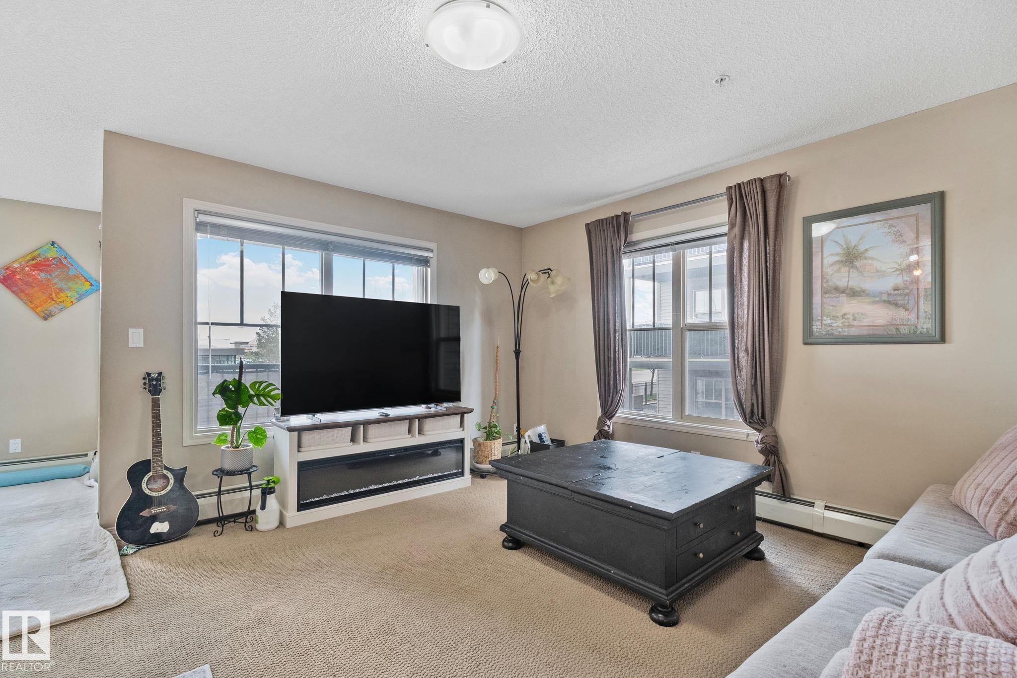 Carpeted living area featuring dual windows with blinds and drapes, a built-in media console with a linear fireplace, and neutral wall tones - 206 103 Ambleside Drive, Edmonton, AB - Indoor