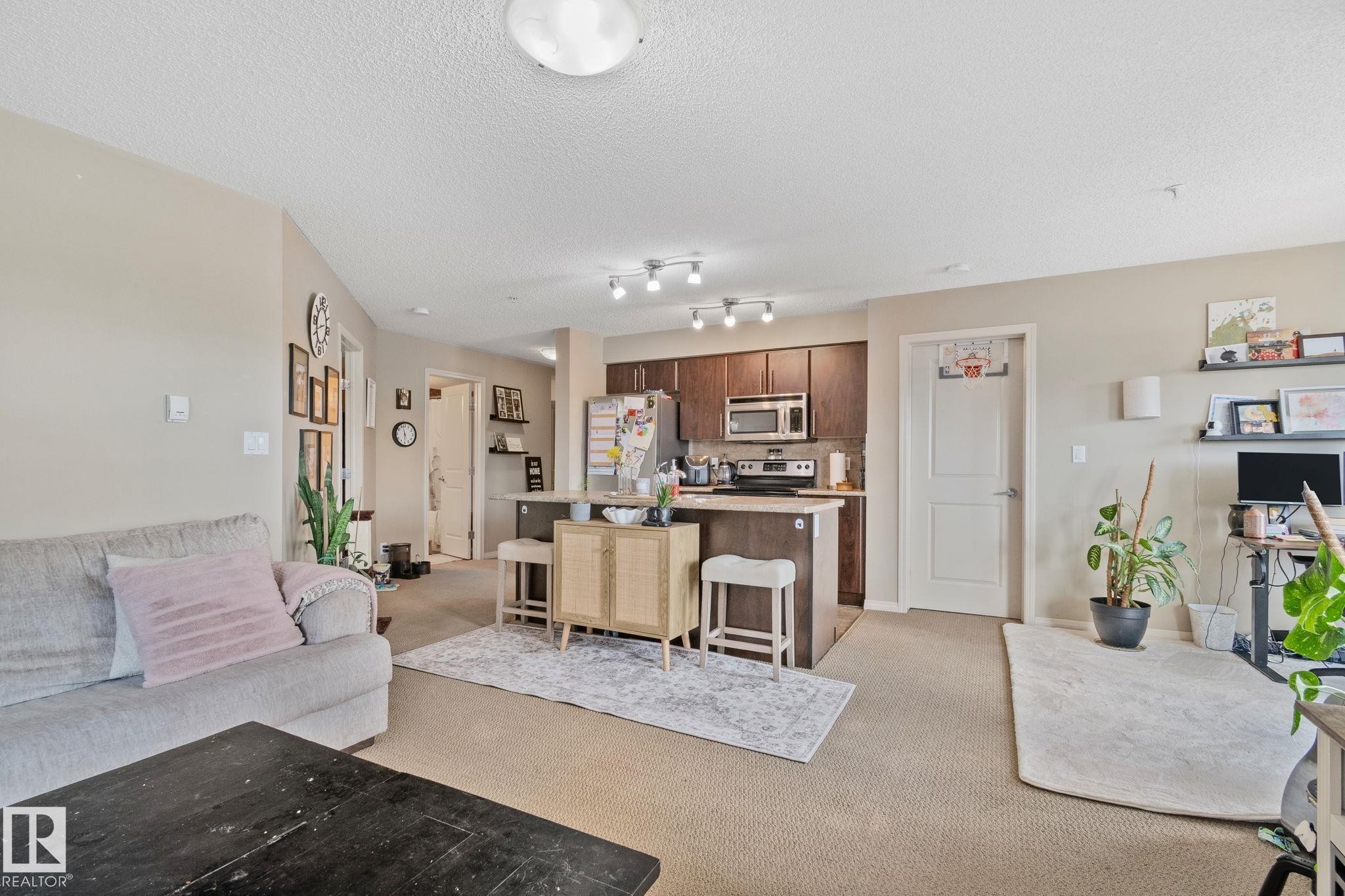 Open-concept living space featuring neutral carpet and paint, recessed ceiling lighting, and a kitchen with dark wood-finish cabinetry, stainless steel appliances, and a breakfast bar - 206 103 Ambleside Drive, Edmonton, AB - Indoor
