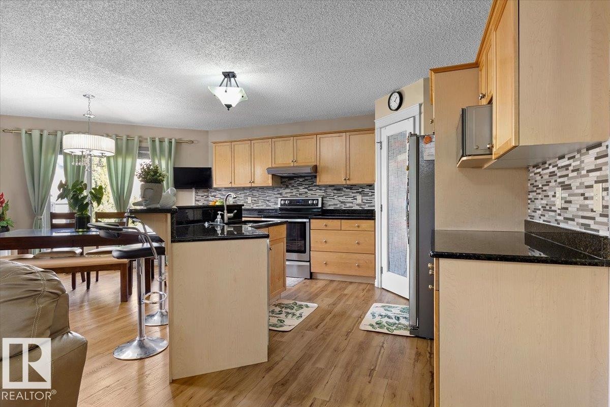 Modern kitchen featuring wood-finish flooring, light wood cabinetry, black countertops, and a mosaic tile backsplash - 3356 25 Ave, Edmonton, AB - Indoor Photo Showing Kitchen